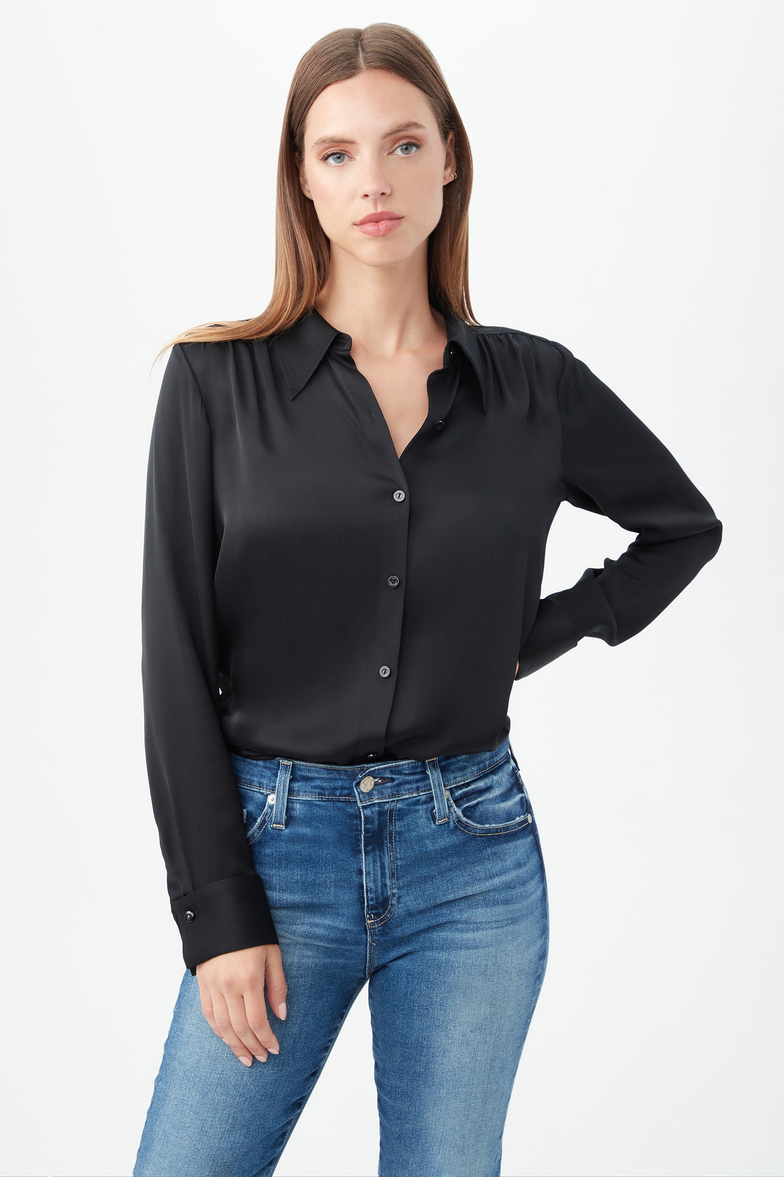 A woman with long, straight brown hair stands against a plain white background, wearing the Cassavetes Long-Sleeve Stretch Silk Button-Up Shirt in black with blue jeans and one hand on her hip.