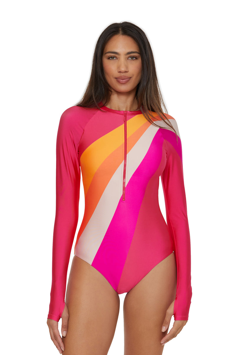 A woman models the Long-Sleeve Zip-Up Paddlesuit in Sunburst—a swimsuit featuring diagonal stripes of pink, orange, yellow, and white—standing against a white background.