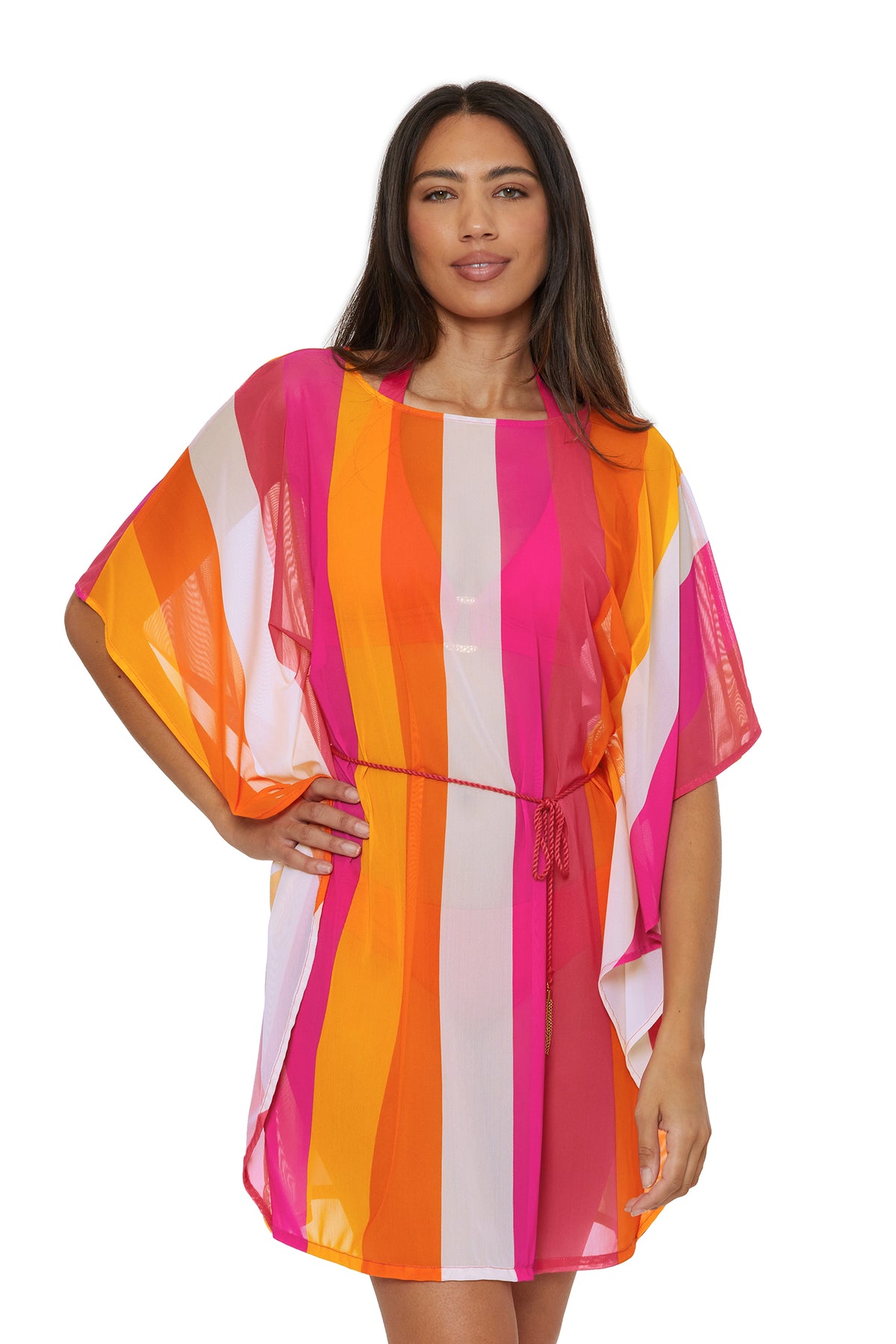 A woman wears the Anissa Cover-Up Caftan in Sunburst—a loose, short-sleeved dress with pink, orange, red, and white stripes and a thin waist belt. She has long straight brown hair and smiles slightly at the camera.