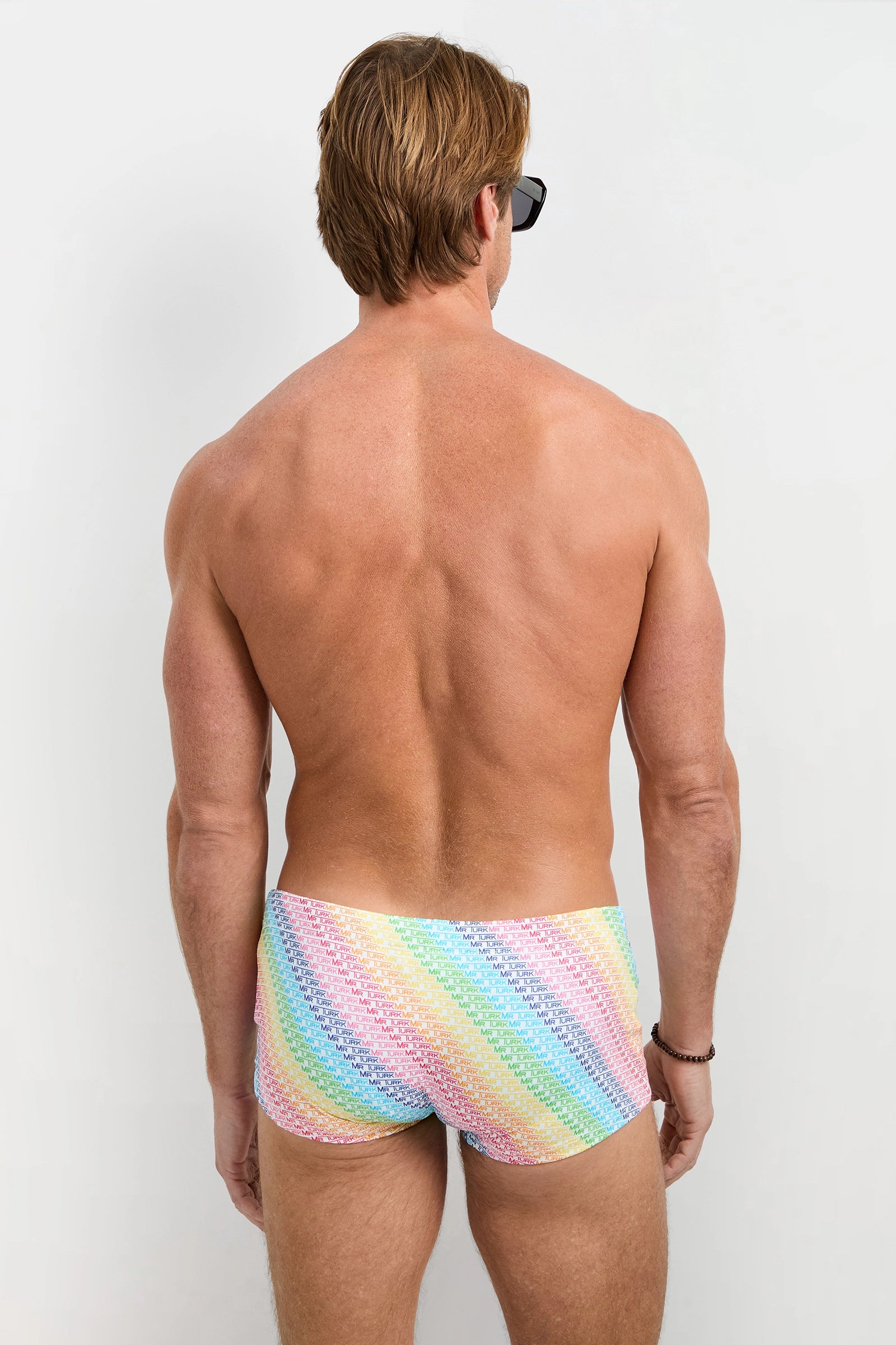 A man with short light brown hair and a tan complexion stands facing away, wearing sunglasses and the Chico Swim Trunk - Pride against a plain light background.