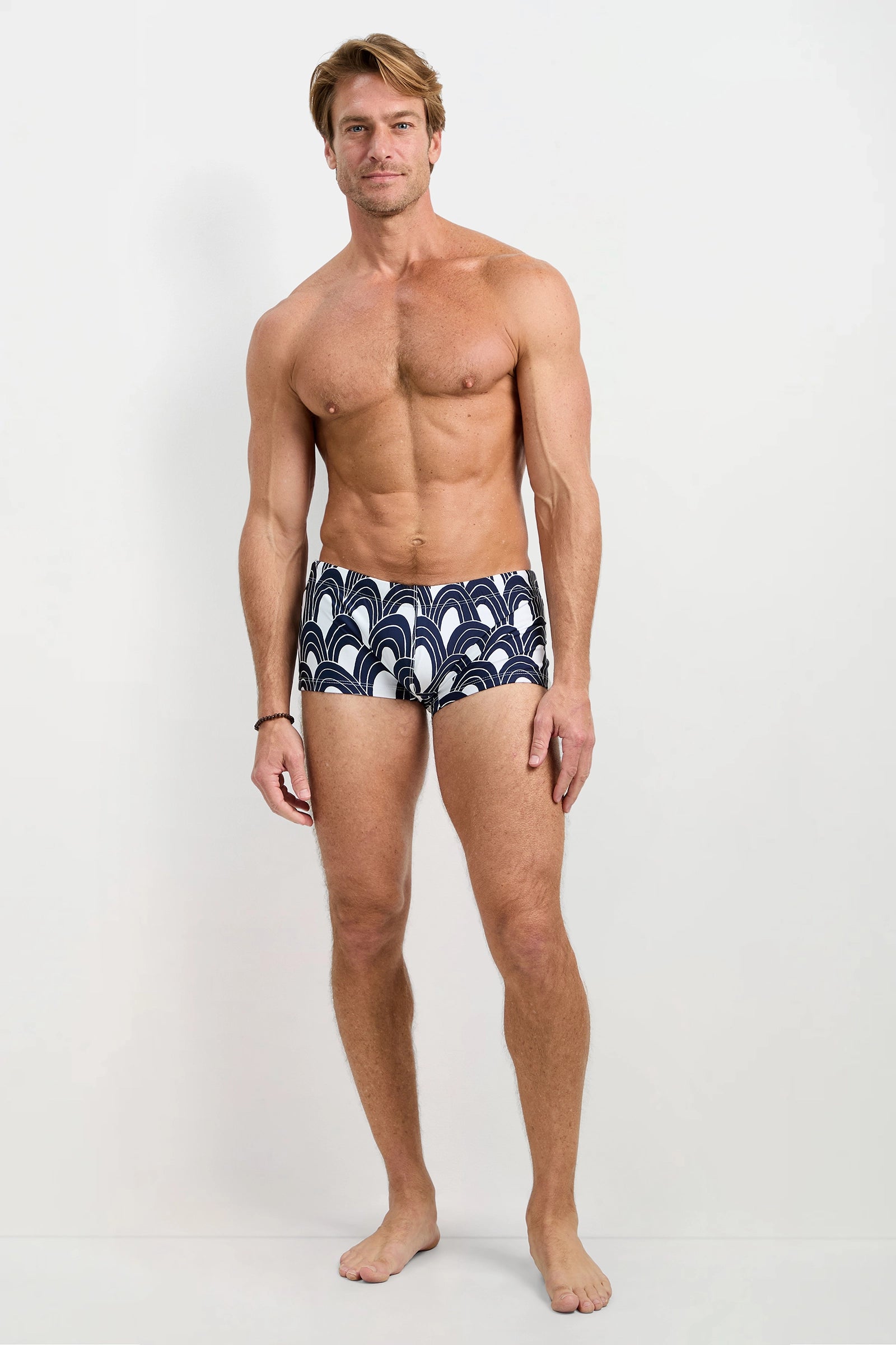 A fit, smiling man with light hair stands barefoot against a plain white background, wearing the Sorrento Swim Trunk in Arches Swim.