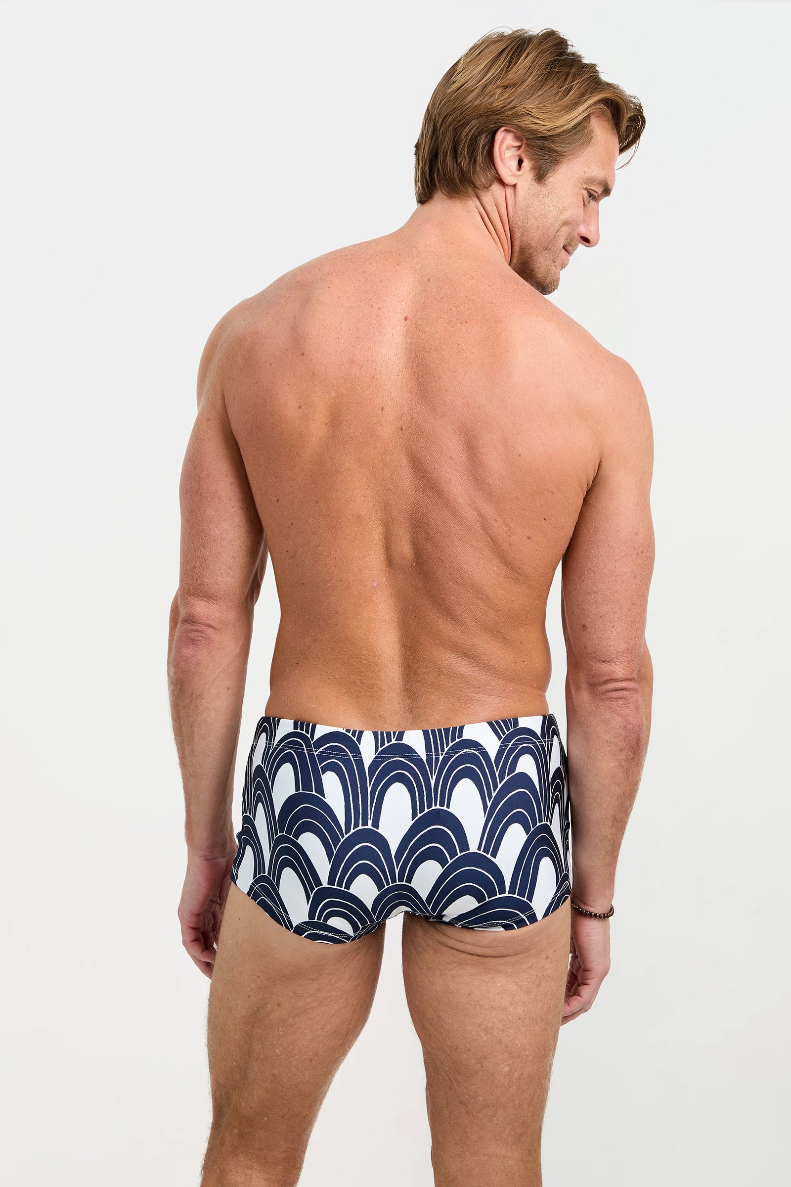 A man with short light brown hair, facing away, wears the Sorrento Swim Trunk - Arches featuring a navy and white abstract arc design, set against a plain light background.
