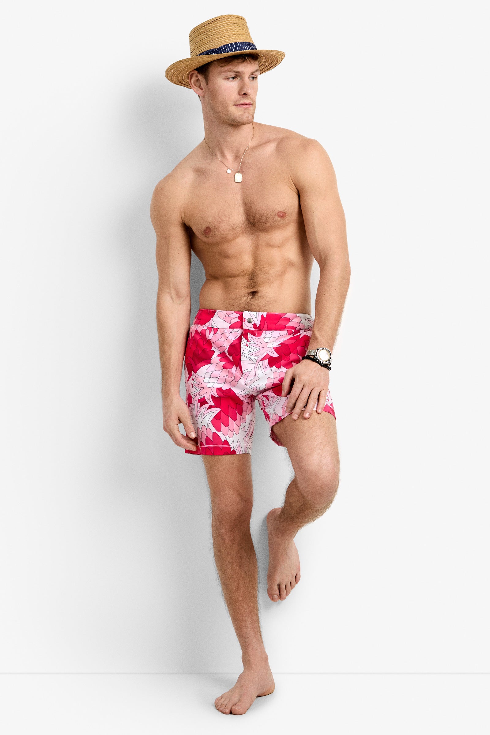 A man stands barefoot against a white background, wearing the Honolulu Board Short—red with a floral pattern—along with a straw hat, necklace, and watch. He looks to the side in a relaxed pose.