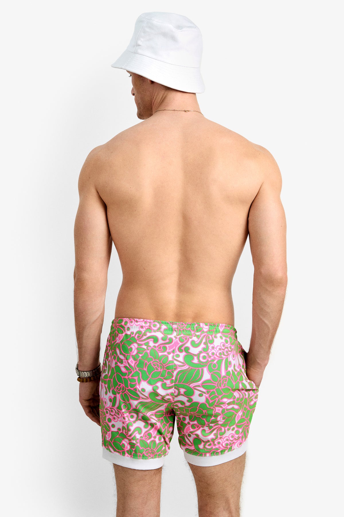 A shirtless man in a white bucket hat and Hermosa Beach Board Short - Petra Floral stands facing away from the camera against a plain white background.