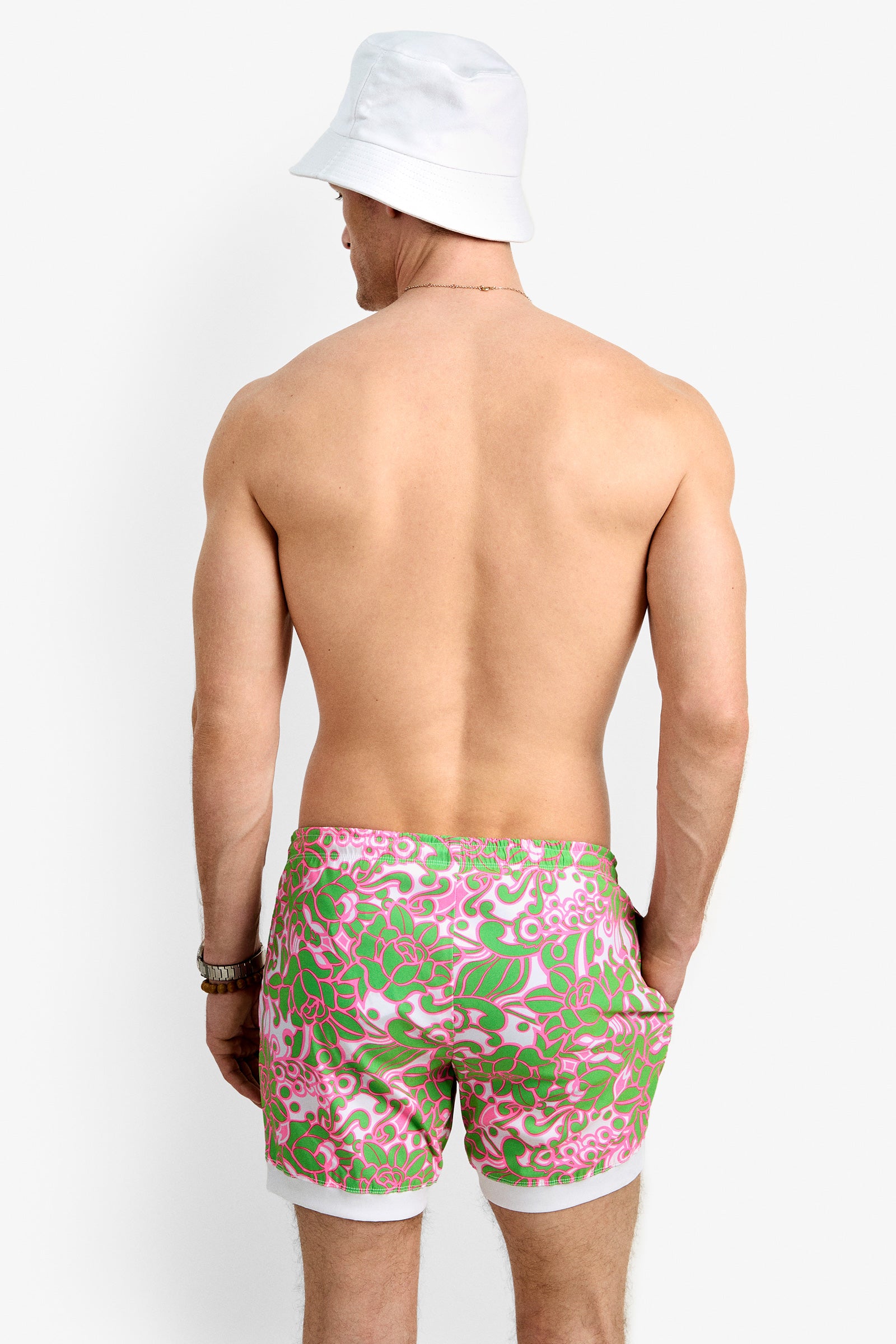 A shirtless man in a white bucket hat and Hermosa Beach Board Short - Petra Floral stands facing away from the camera against a plain white background.