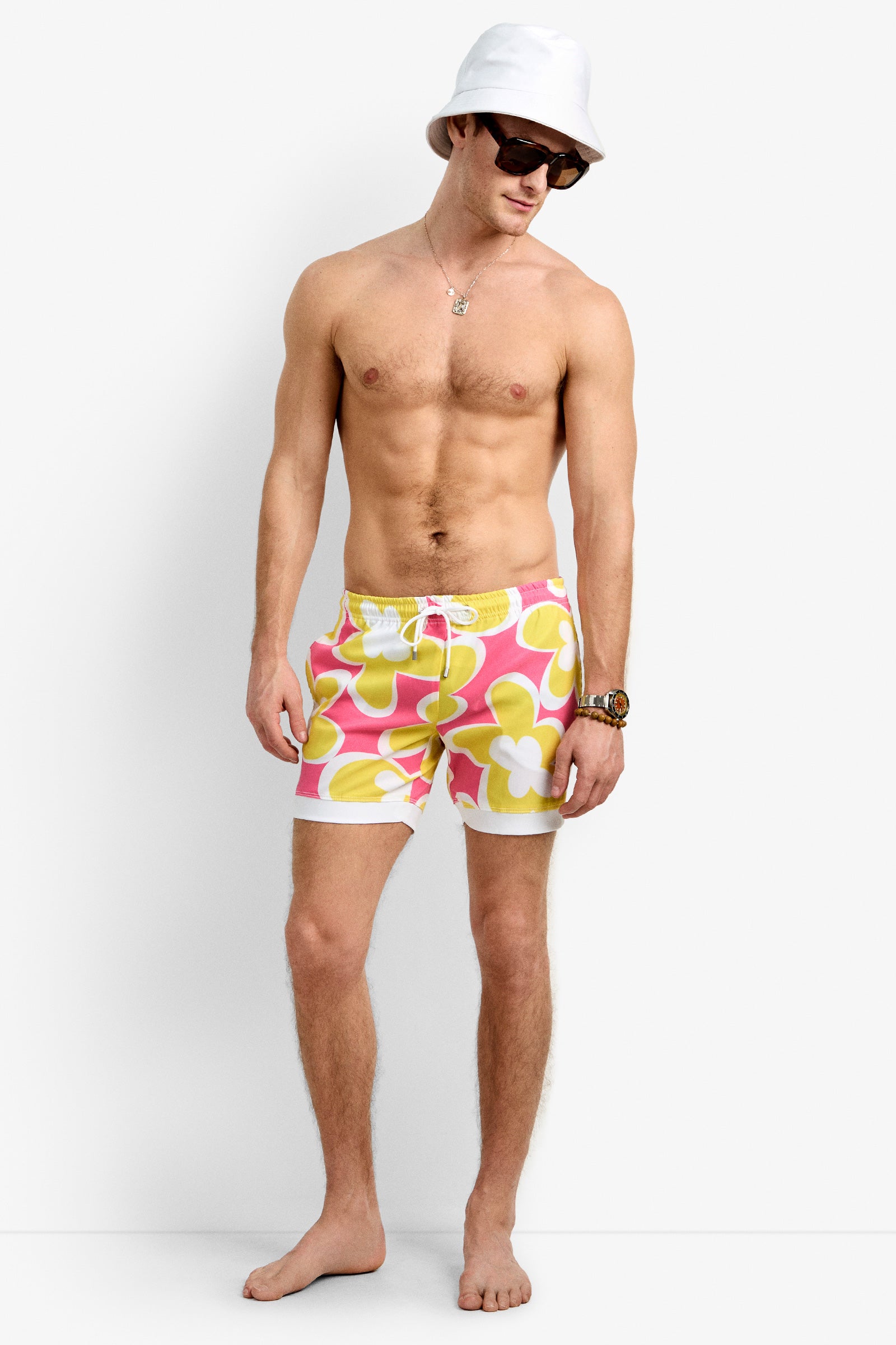 A man wearing a white bucket hat, sunglasses, and Hermosa Beach Board Short swimsuits with pink, yellow, and white floral patterns stands barefoot against a plain white background.