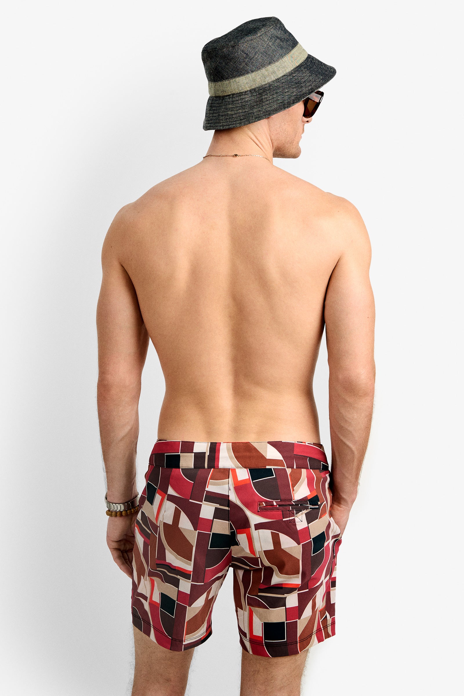 A shirtless man faces away in a dark bucket hat, sunglasses, bracelet, necklace, and the Honolulu Board Short—patterned red and brown swim trunks—against a plain light background.