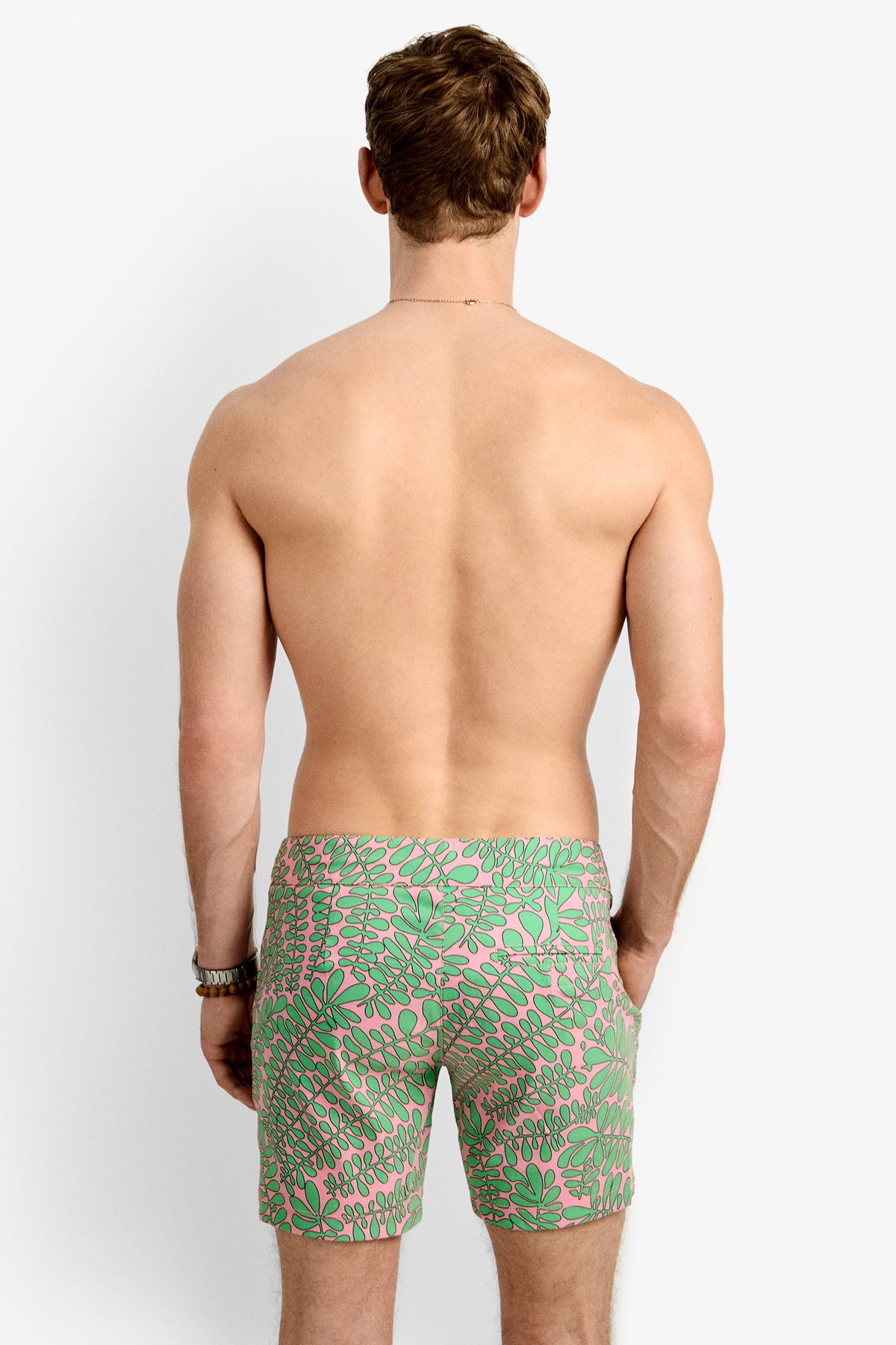 A shirtless man stands with his back to the camera, modeling the Honolulu Board Short - Vines, which features a green and pink leafy pattern. The background is plain white.