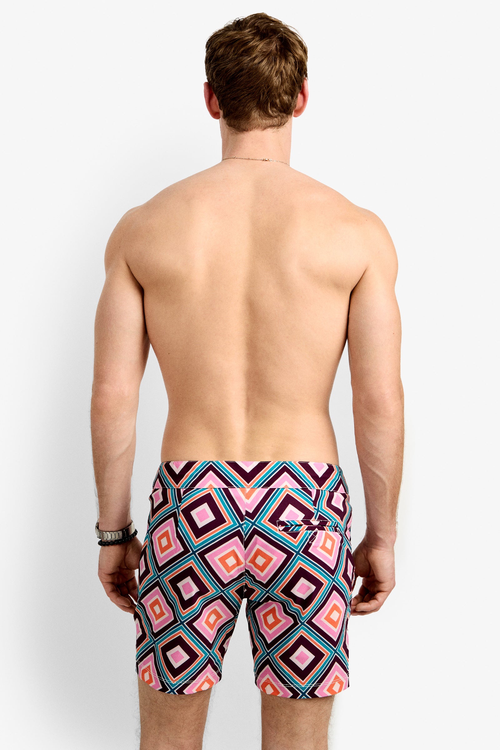 A man with short brown hair is seen from behind, shirtless and wearing the Honolulu Board Short featuring a colorful geometric pattern in pink, red, blue, and black.