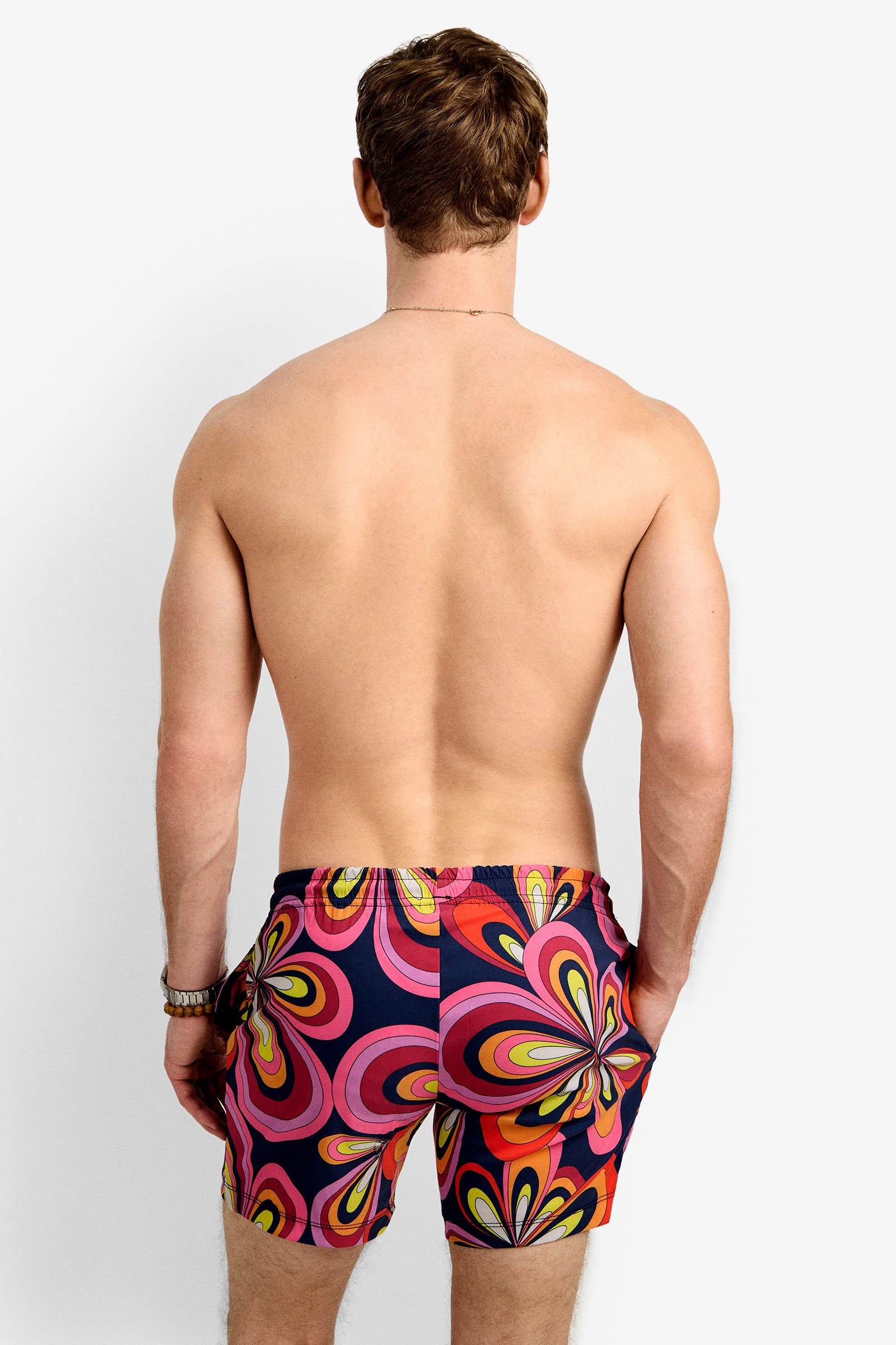 A man with short brown hair, shirtless and facing away, wears the Imperial Beach Swim Trunk - Boca Blooms featuring a bold, retro floral pattern in pink, yellow, orange, and navy blue.