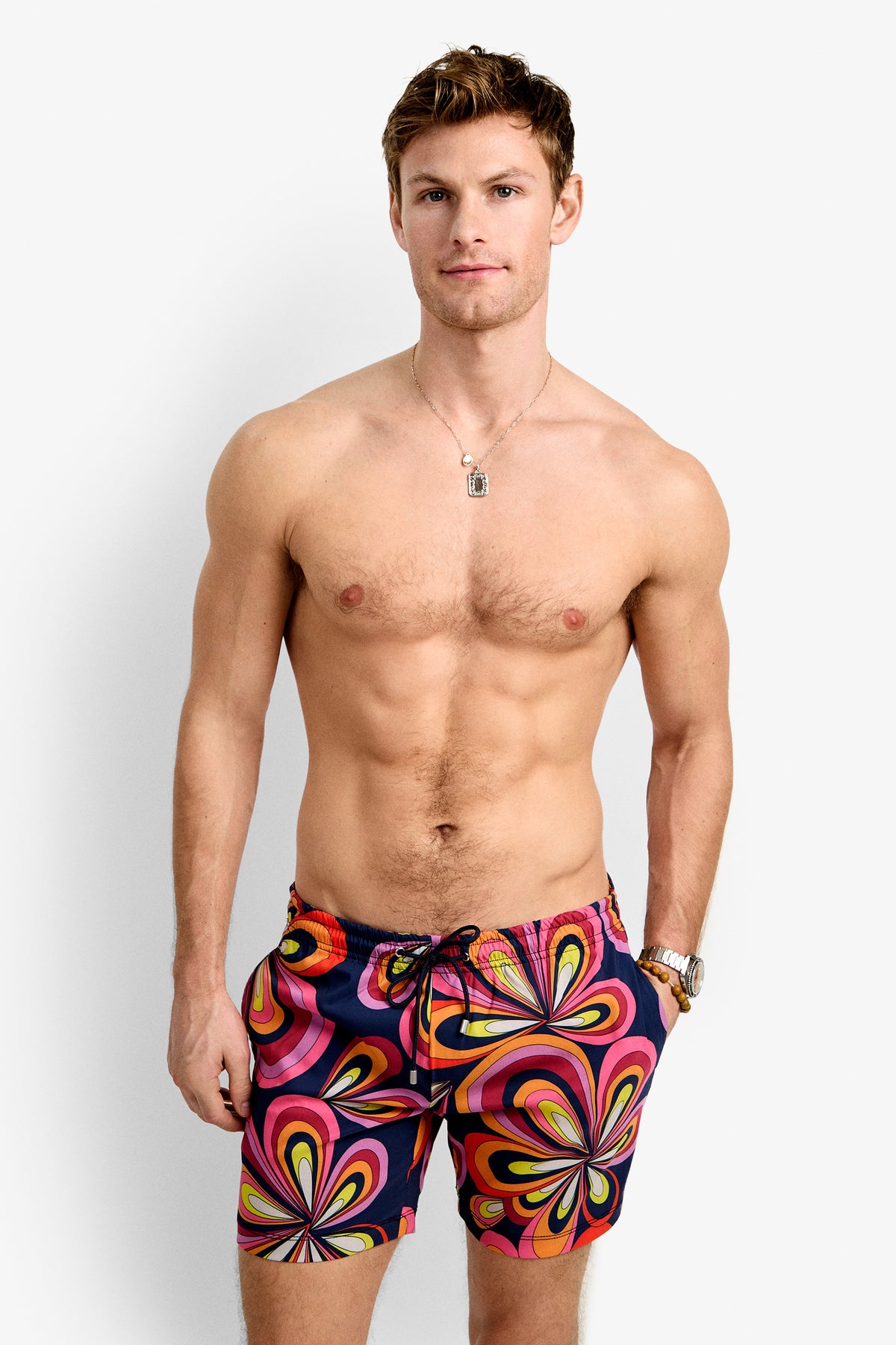A man with short brown hair models the Imperial Beach Swim Trunk - Boca Blooms, featuring a colorful retro pattern and drawstring, against a plain white background. He is shirtless and accessorized with a necklace and wristwatch.