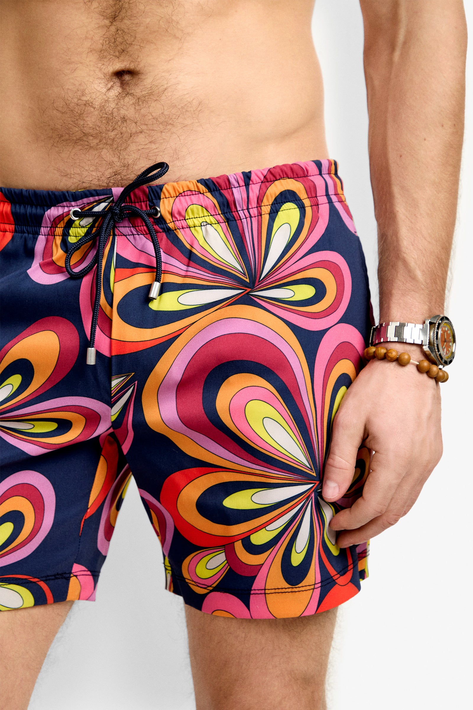 A man wears the Imperial Beach Swim Trunk – Boca Blooms, featuring colorful retro swirls of pink, orange, yellow, and purple. He also sports a beaded bracelet and a watch; only his torso and upper legs are visible.