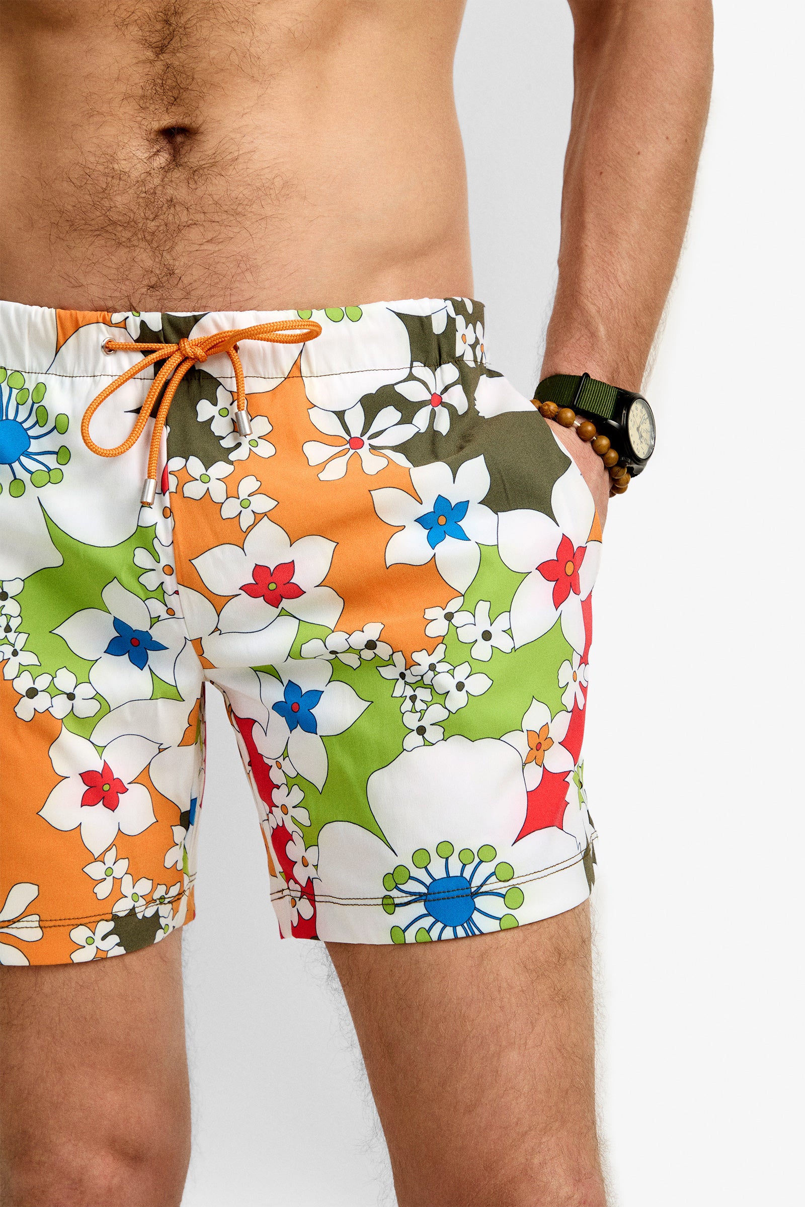 A person stands with one hand in the pocket, wearing Imperial Beach Swim Trunks featuring colorful floral patterns of orange, green, and white. They also wear a beaded bracelet and a wristwatch; the upper body is partially visible.