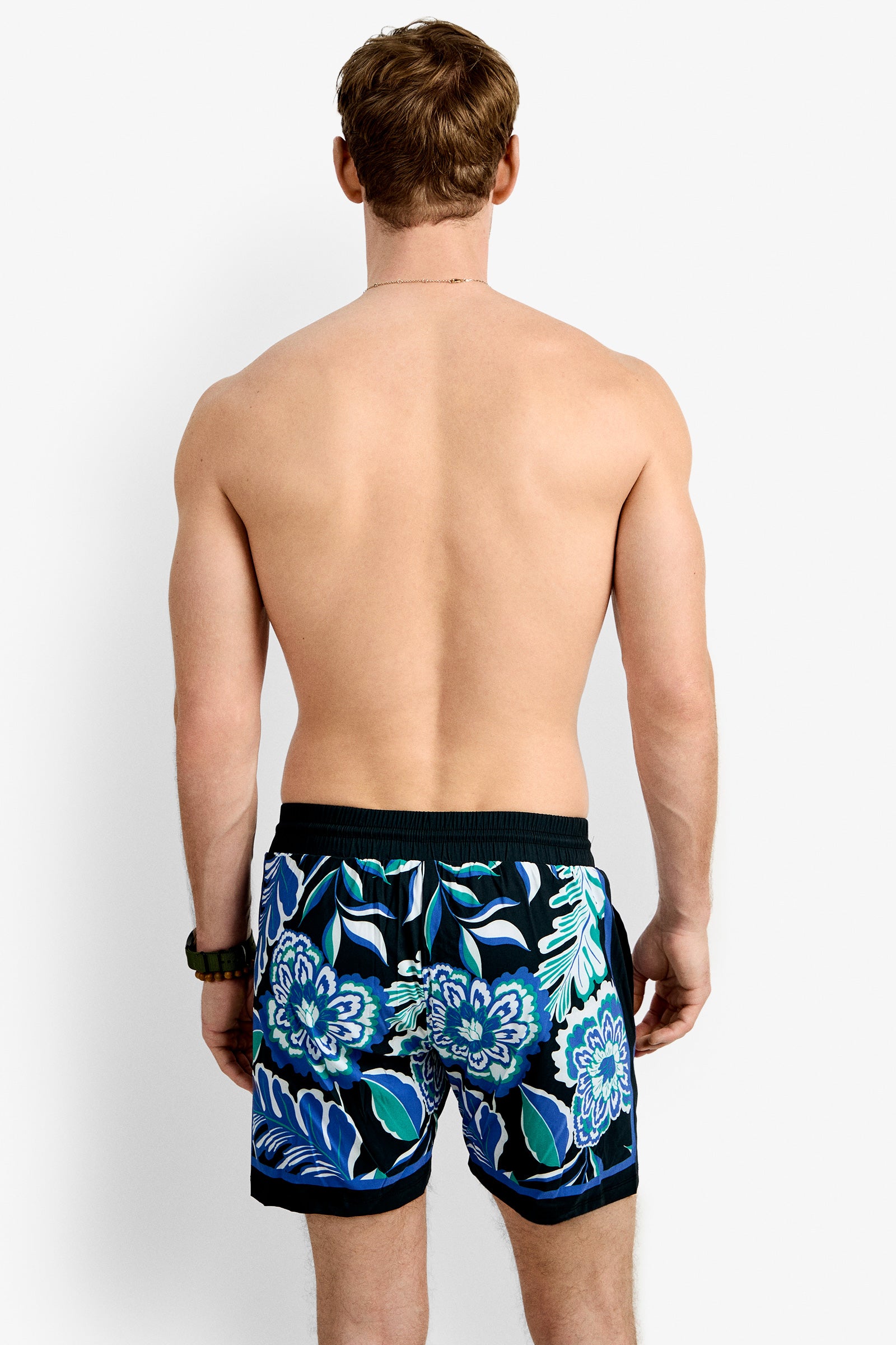 A man with light skin and short brown hair, shirtless and facing away, models Bruce's Beach Swim Trunk featuring a blue, green, and white floral pattern. The background is plain white.