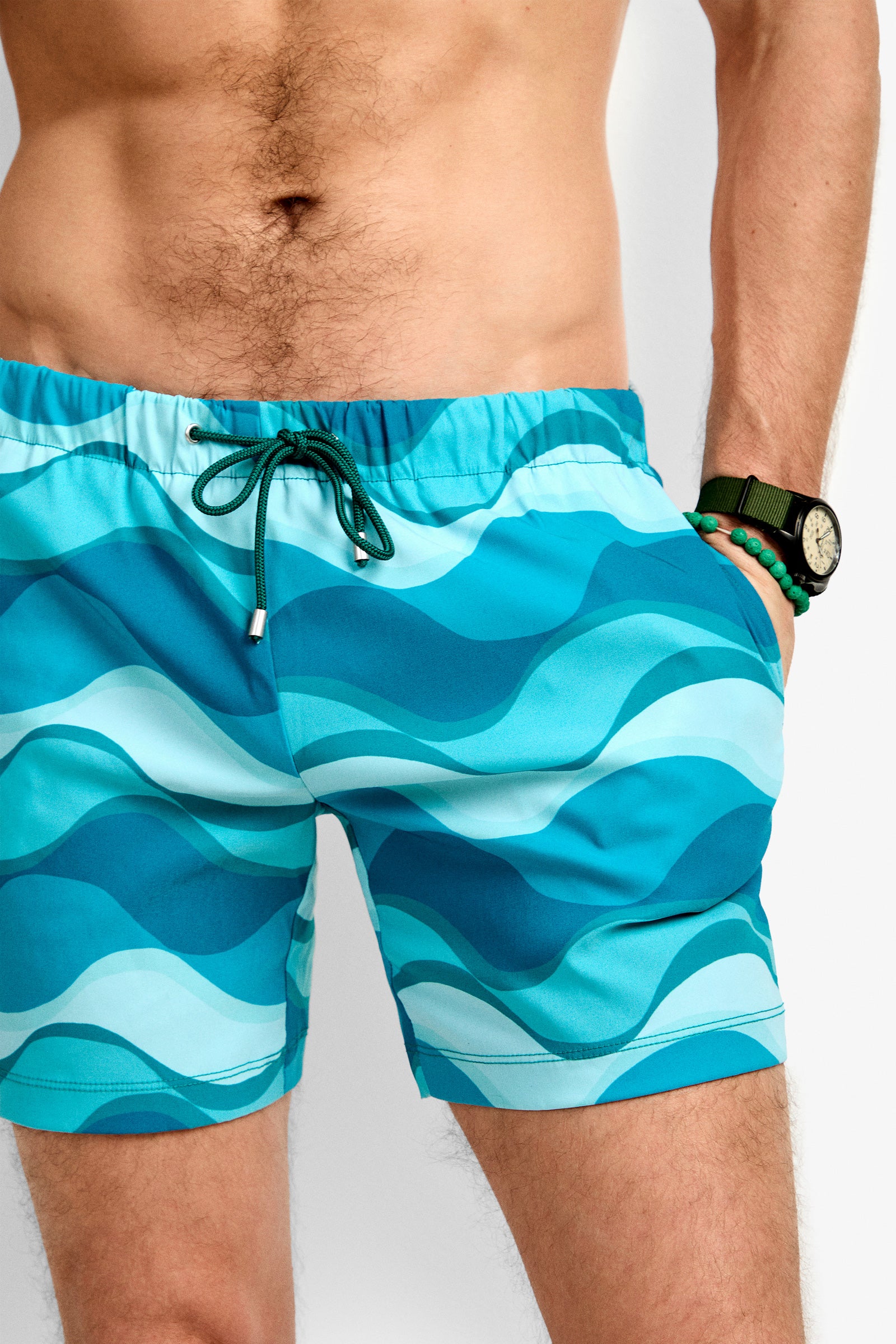 A man wearing the Imperial Beach Swim Trunk, featuring blue wave patterns, stands with one hand in his pocket. He sports a watch and a green beaded bracelet on his bare wrist.
