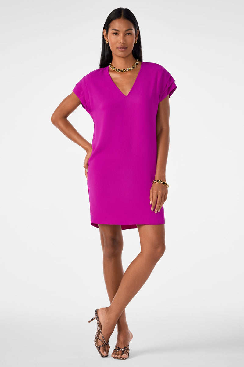 A woman stands confidently against a plain white background, wearing the Sadora Short-Sleeve Dress in Geranium, accessorized with gold jewelry and open-toed snakeskin heels.