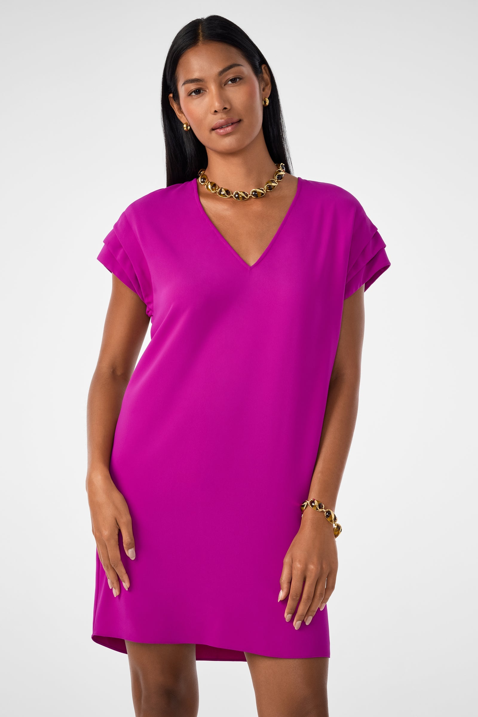 A woman wears the Sadora Short-Sleeve Dress in Geranium, featuring a V-neck and layered short sleeves, styled with a chunky gold necklace and bracelet. She stands against a white background with straight dark hair and a relaxed expression.