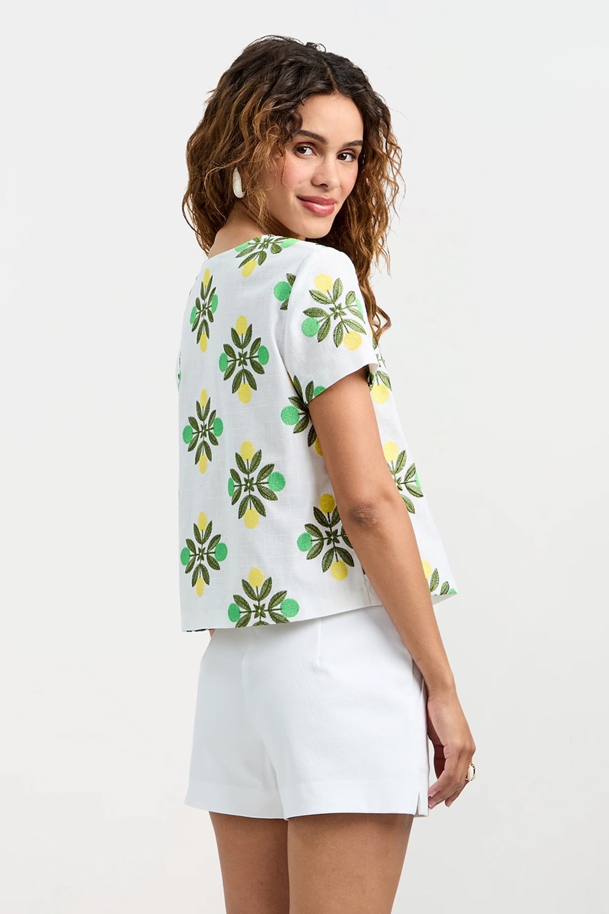 A woman with wavy hair wears the Salina Tie-Front Top, a white short-sleeve shirt featuring green and yellow floral patterns, paired with white shorts. She stands sideways, glancing over her shoulder with a slight smile.