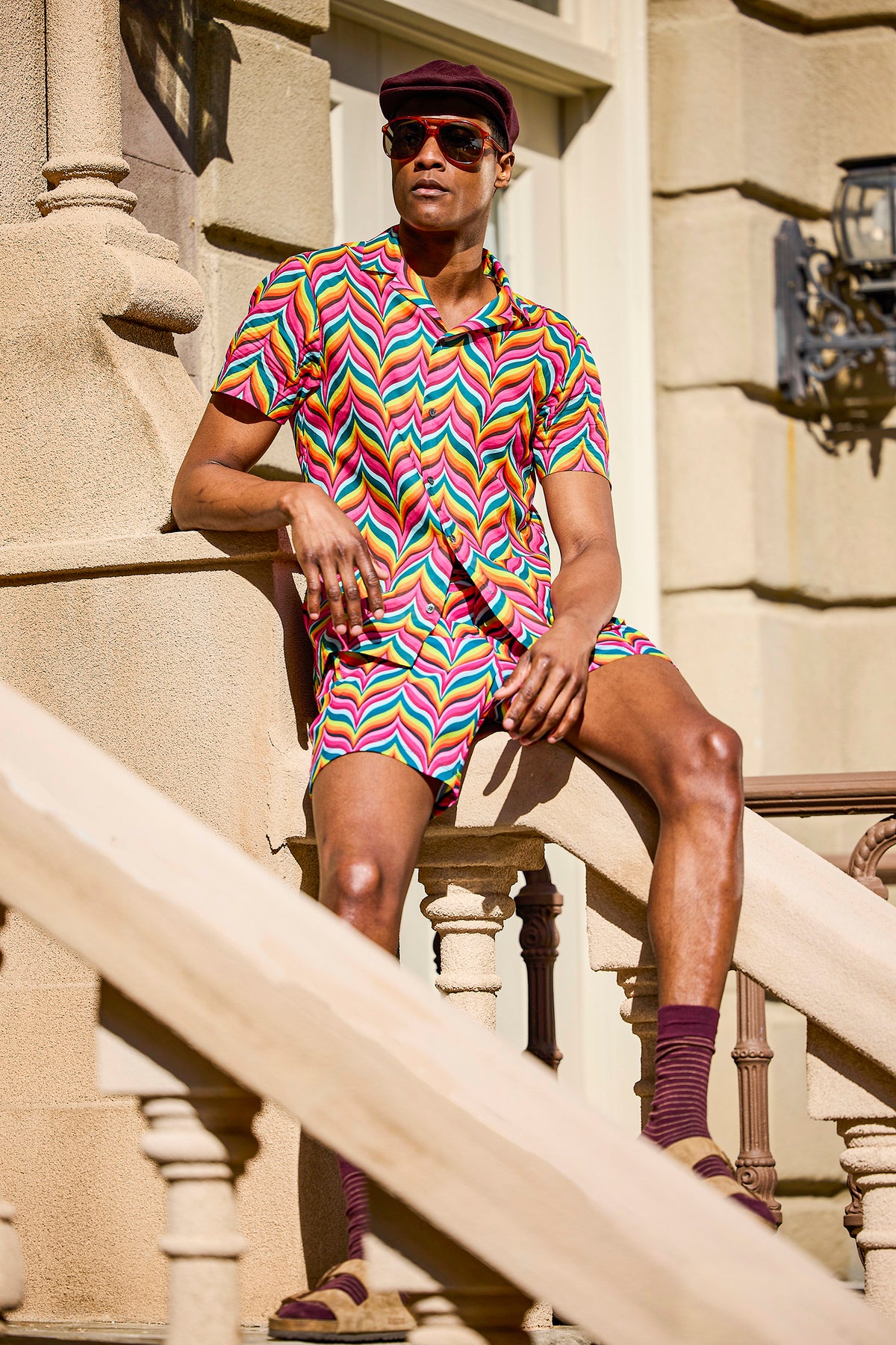 A person sits on outdoor stairs wearing the Felix Shirt—an organic cotton camp shirt with a colorful zigzag pattern—and matching shorts, plus sunglasses, a maroon cap and socks, beige sandals, with a stone building behind them.