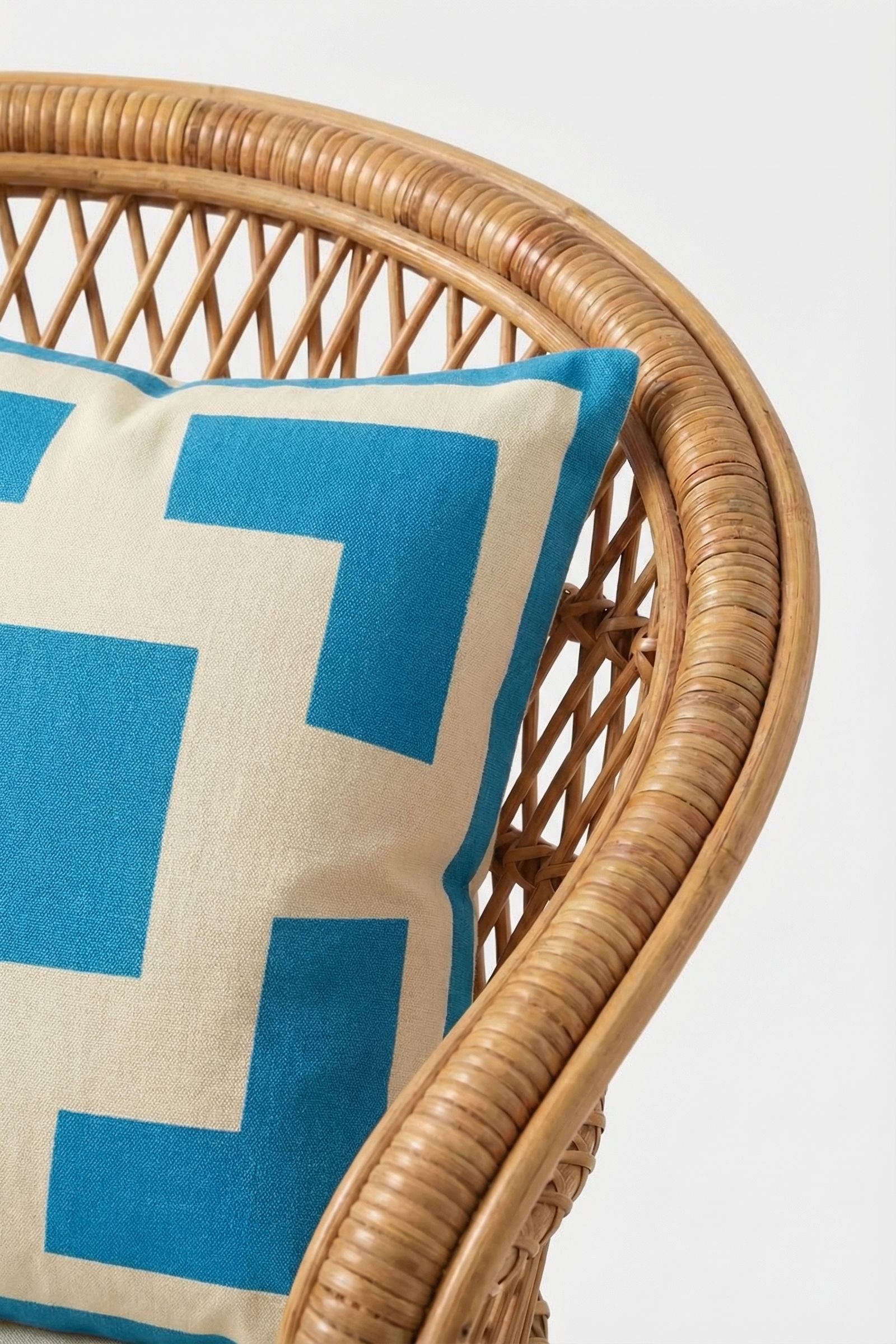 A close-up of a wicker chair with the Palm Springs Block Embroidered Pillow, 20"x20" and down-filled, featuring a geometric pattern against a plain white background.