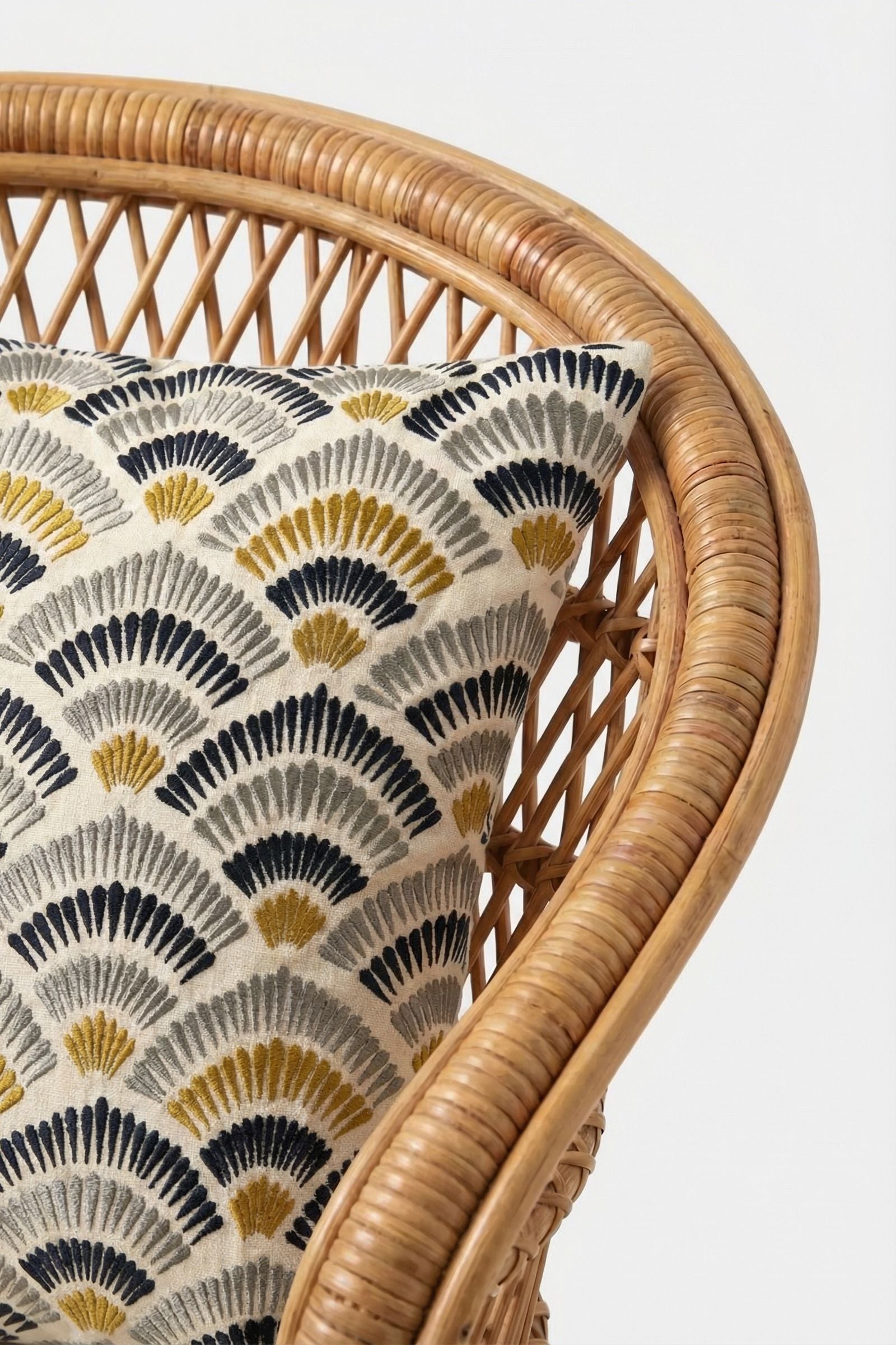 A close-up of a woven rattan chair with the Pasadena Embroidered Square Pillow, showcasing a repeating fan pattern in black, grey, and yellow on a cream background.