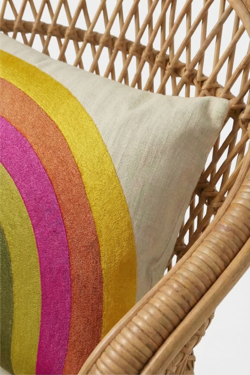 A close-up of a woven rattan chair with a cream cushion featuring curved yellow, orange, pink, and green stripes. The Calistoga Embroidered Lumbar Pillow adds an extra pop of color. The background is white.