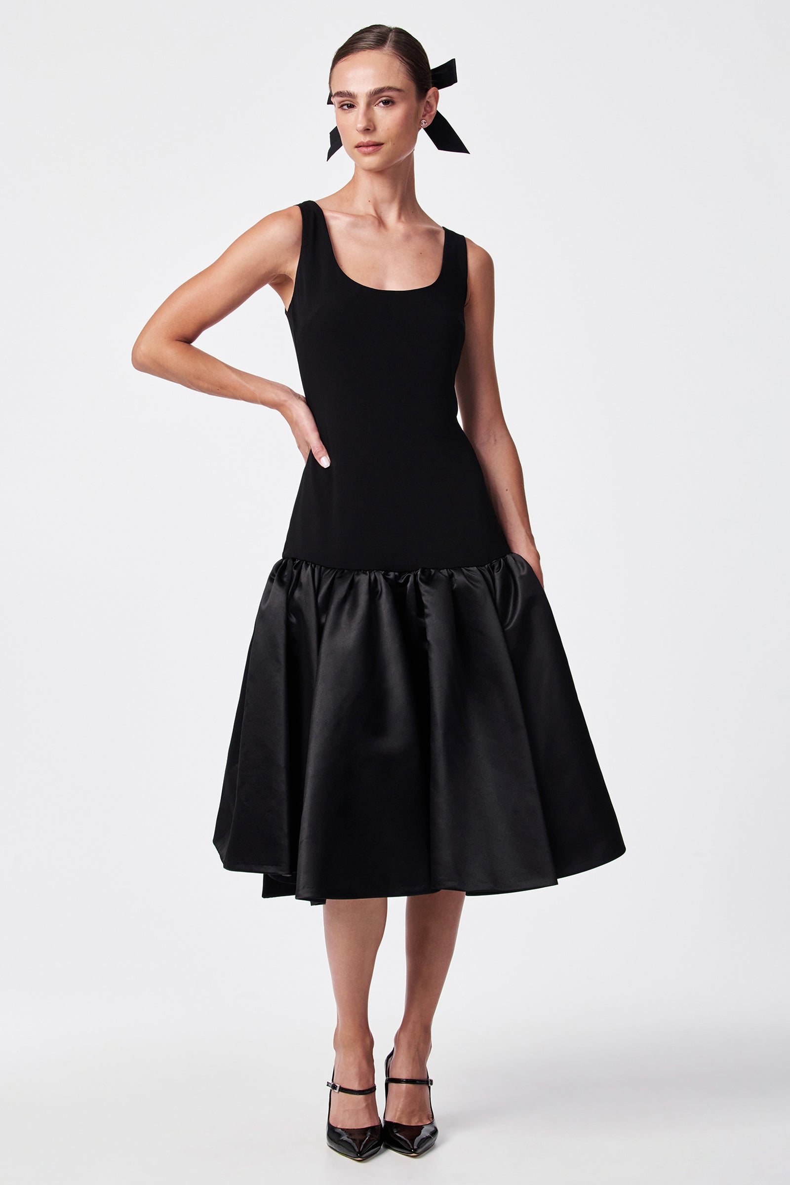 A woman models the SINA DRESS - BLACK, featuring a sleeveless fitted bodice and flared satin skirt. She accessorizes with a black ribbon in her hair and black heels against a plain white background.