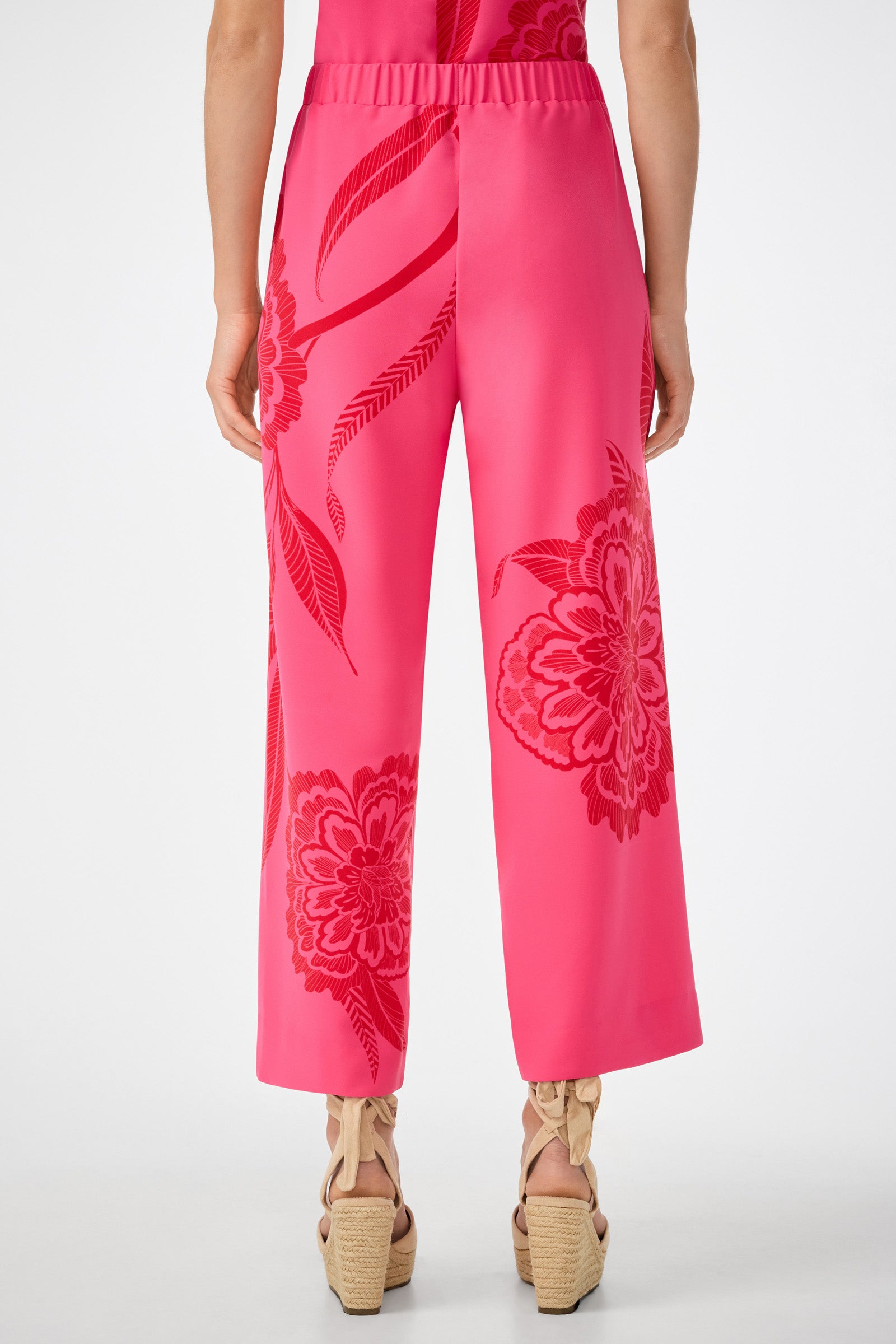 Someone standing with their back to the camera is wearing the Statue Pant in bright pink with bold red floral patterns, paired with beige platform sandals.