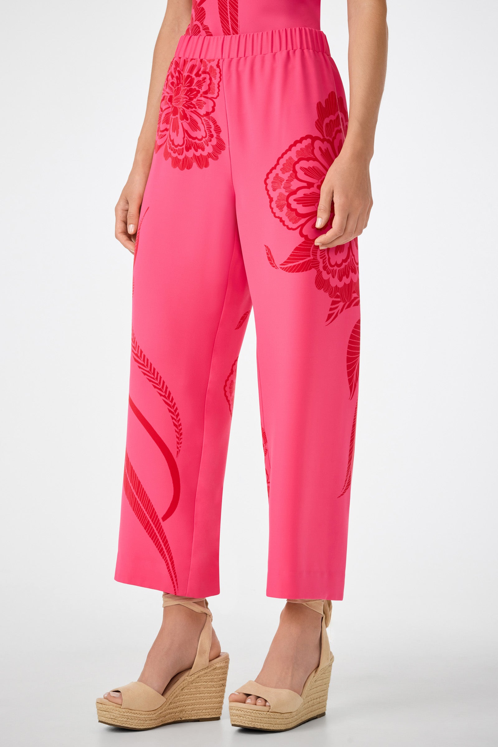 Someone wearing the Statue Pant in bright pink with a floral pattern and wide legs, paired with beige wedge sandals. The image captures the outfit from mid-torso to feet against a plain background.