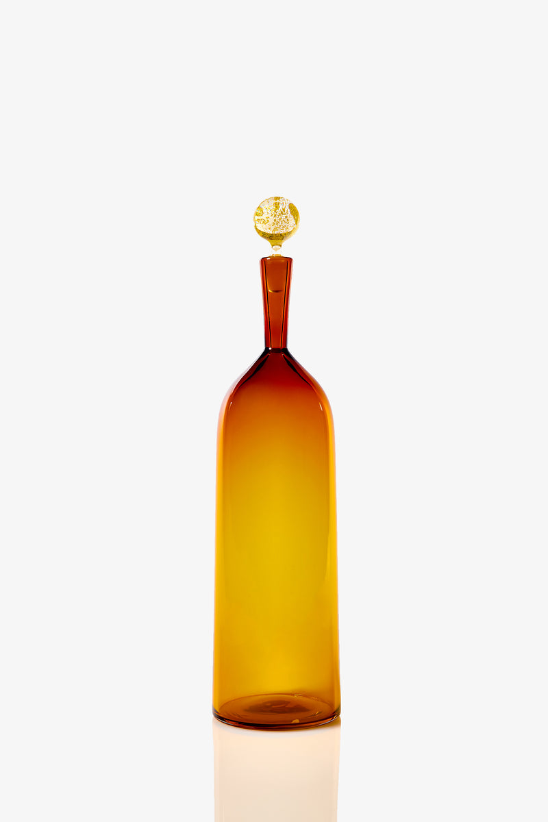The Tall Bottle Carmella Barware is a tall, cylindrical amber glass bottle with a narrow neck and decorative round stopper, displayed against a plain white background.