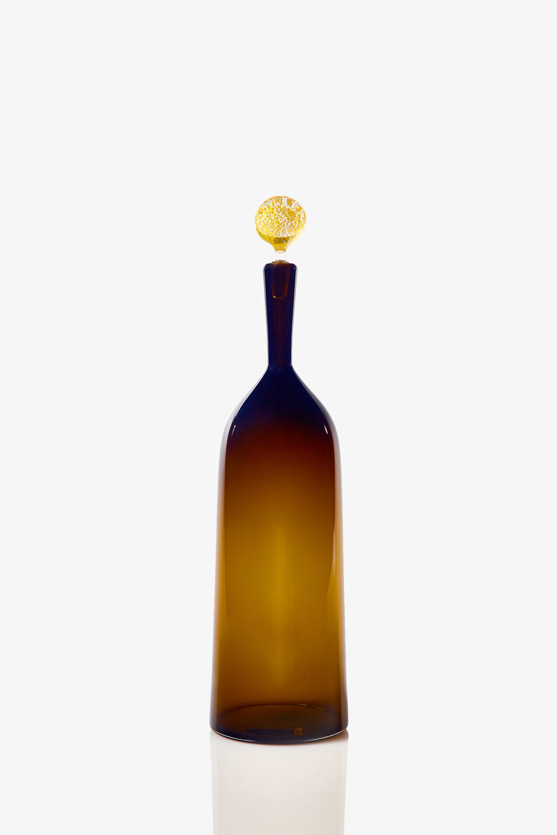 The Tall Bottle Carmella Barware features a tall, amber-colored glass body with a narrow neck and a round, textured golden stopper, showcased against a plain white background.