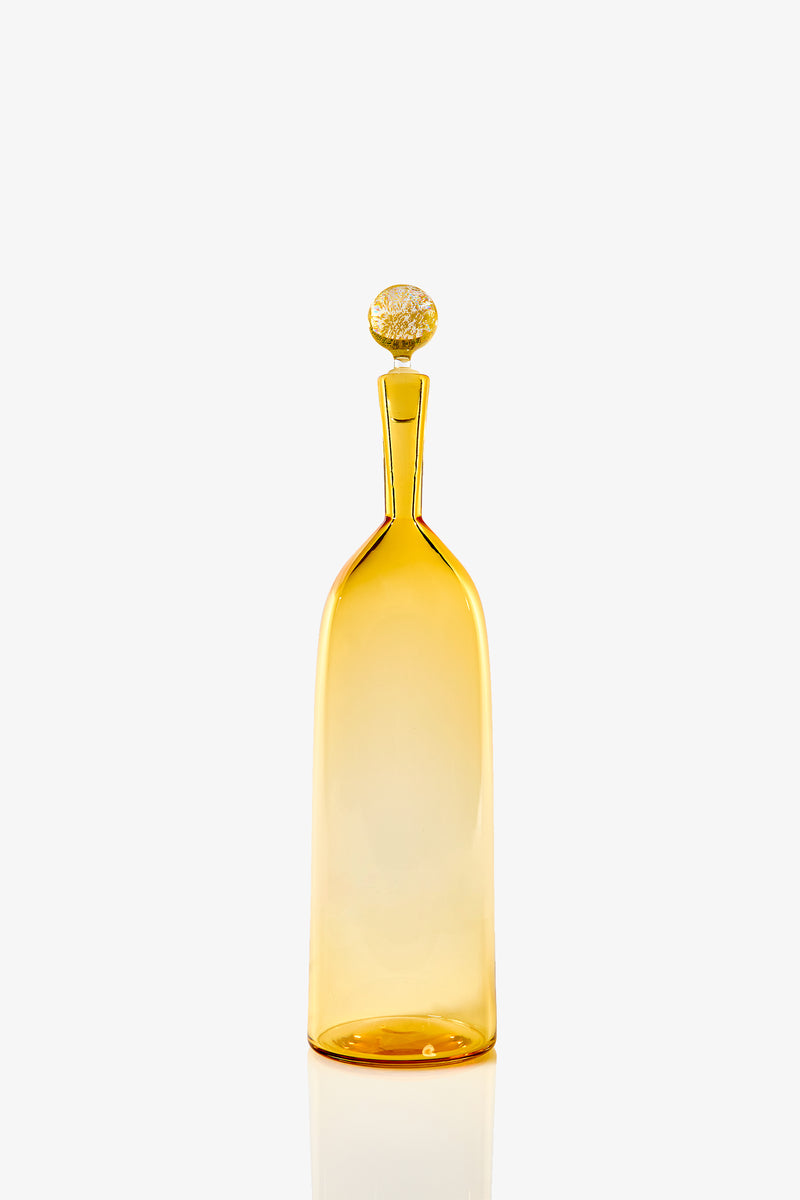 The Tall Bottle Carmella Barware is a slender amber glass bottle with a narrow neck and round, textured stopper, shown against a plain white background.