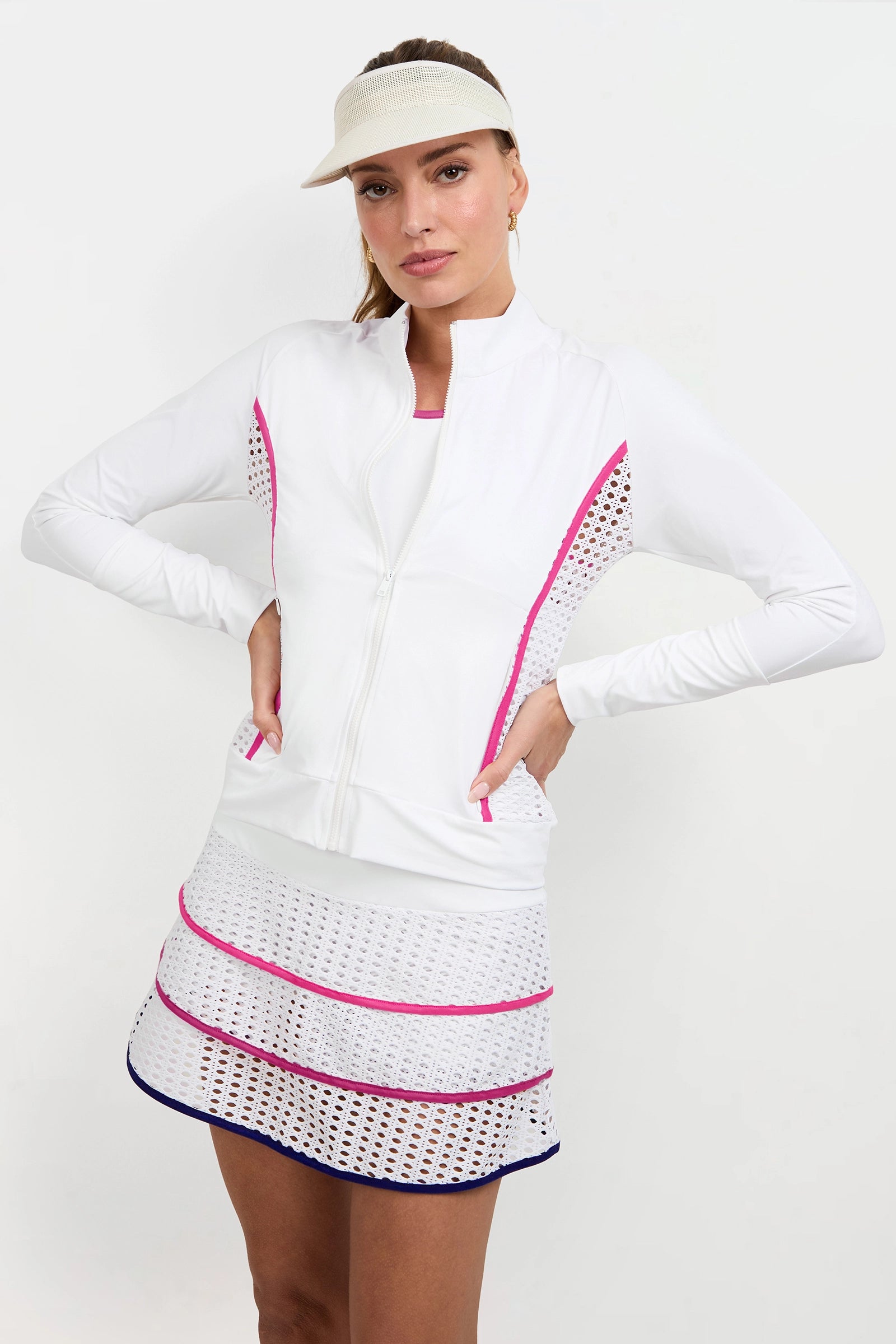 A woman stands with her hands on her hips against a plain white background, wearing a Performance Jacket—white with pink accents—a white visor, and a matching skirt featuring mesh panels and pink and navy stripes.