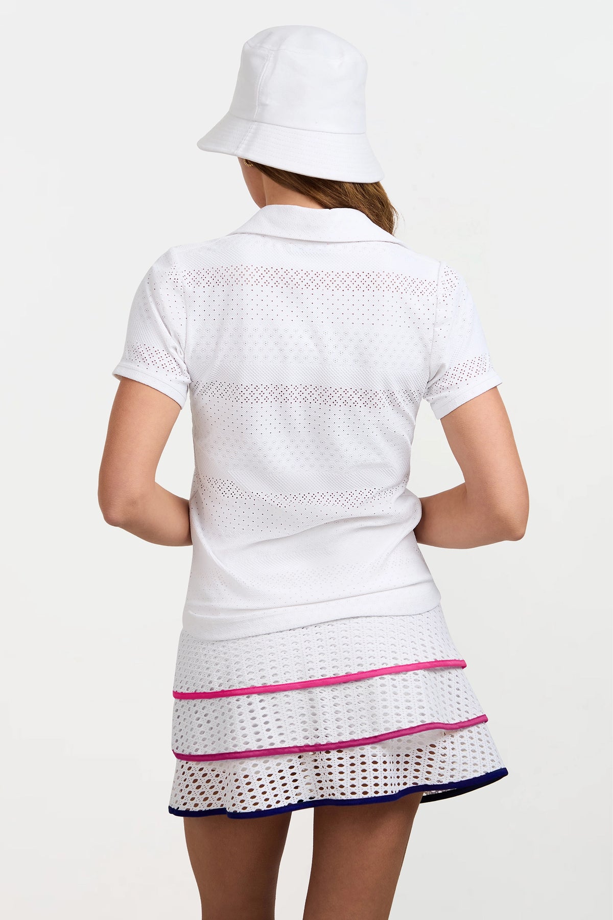 A woman seen from behind wears a white bucket hat and the Lace Polo, a white perforated short-sleeve top, paired with a matching skirt featuring horizontal pink and dark blue stripes near the hem.
