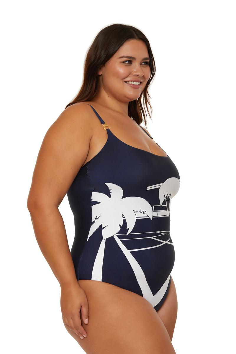 A woman with long brown hair smiles while wearing the Scoop Neck One-Piece Swimsuit - Trina Tennis, featuring a navy blue color and white tropical beach design with palm trees, as she poses against a plain white background.