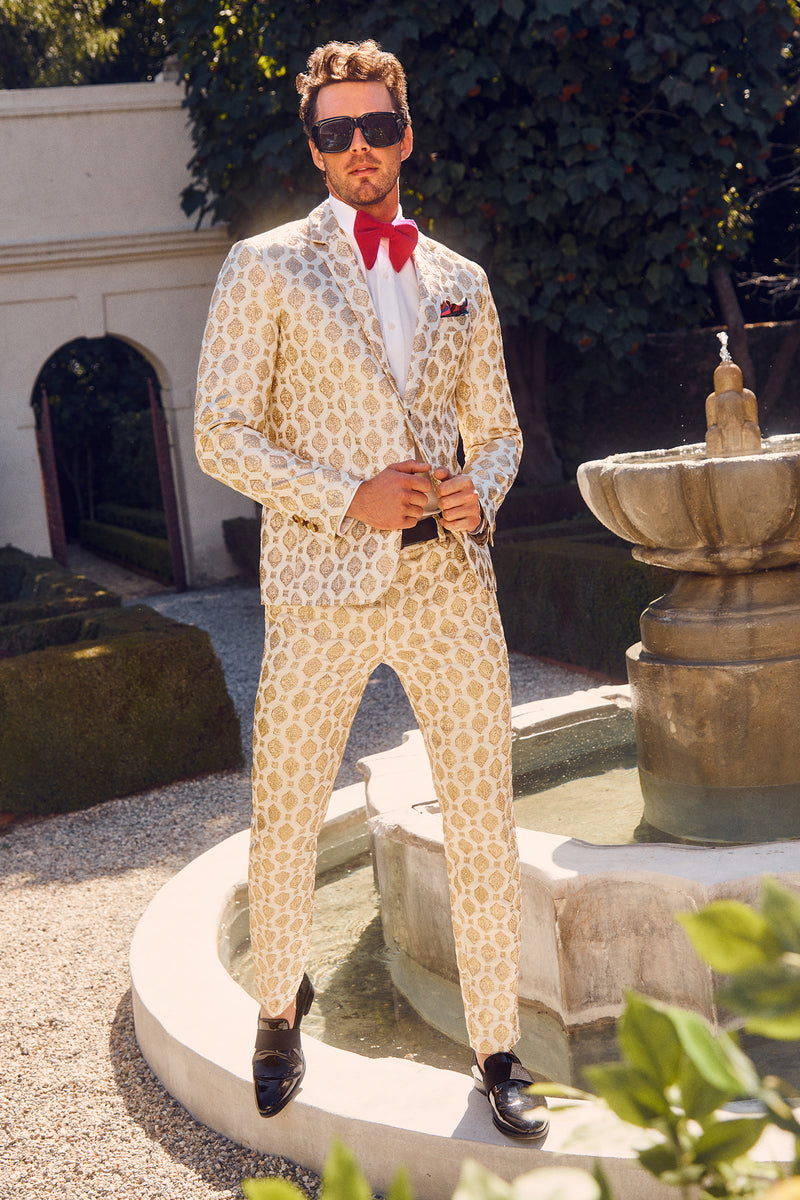 A man stands confidently by a stone fountain in a sunlit garden, wearing the Clyde Slim Trouser - Jubilant Jacquard in Winter White with a matching jacket, red bow tie and pocket square, black loafers, and large sunglasses.