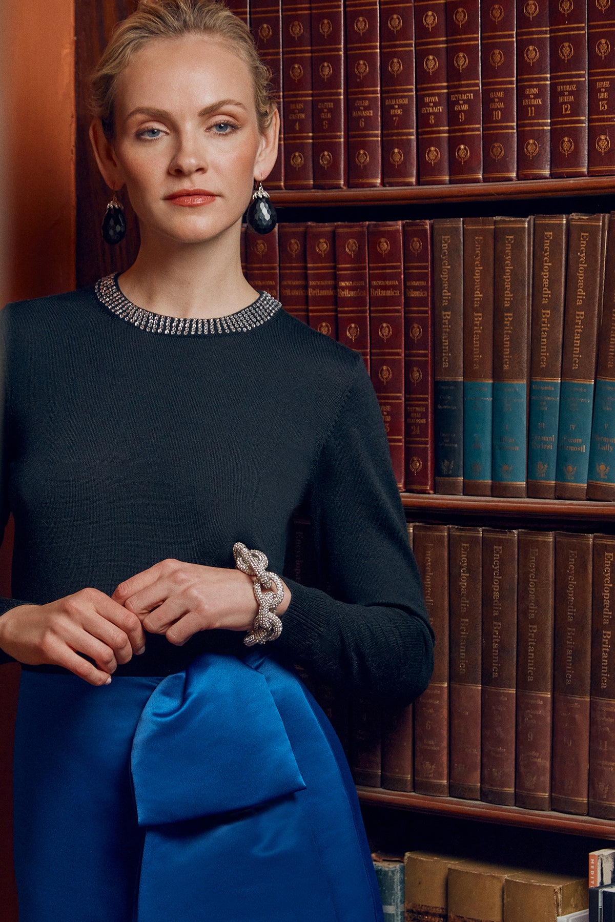 A woman wearing the Laelia Merino Wool Sweater with Rhinestone Trim stands before a bookshelf of leather-bound books, paired with statement earrings, a silver bracelet, and a blue skirt featuring a large bow.