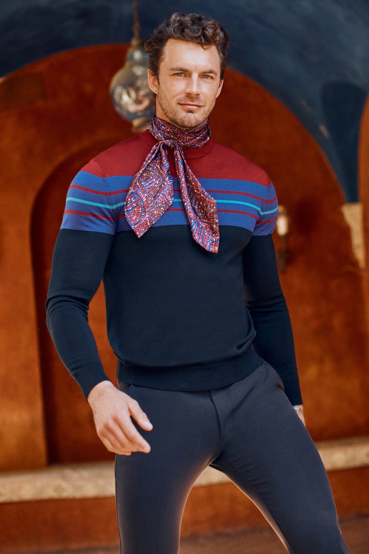 A man poses confidently against a warm terracotta backdrop, wearing the Romero Crewneck Sweaterâ€” a fitted, long-sleeve color-block design with horizontal stripesâ€”paired with a patterned red scarf.