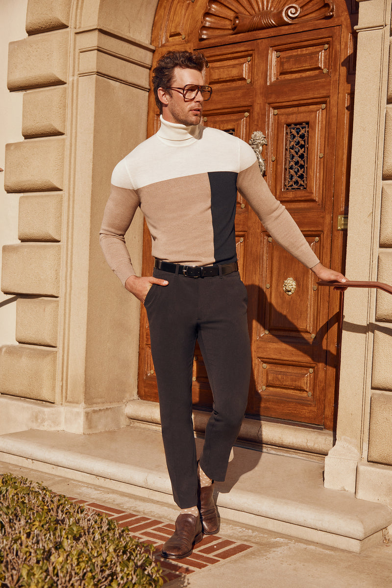 A man wearing glasses and the Callisto Turtleneck Sweater - Merino Wool in beige, white, and black stands confidently with one hand in his pocket before an ornate wooden door, paired with dark pants and brown shoes.