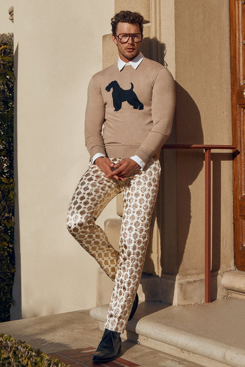 A man in glasses wears the Weston Airedale Terrier Sweaterâ€”crafted from merino wool with a black dog silhouetteâ€”paired with patterned cream and gold pants, standing on stone steps in sunlight by a building.