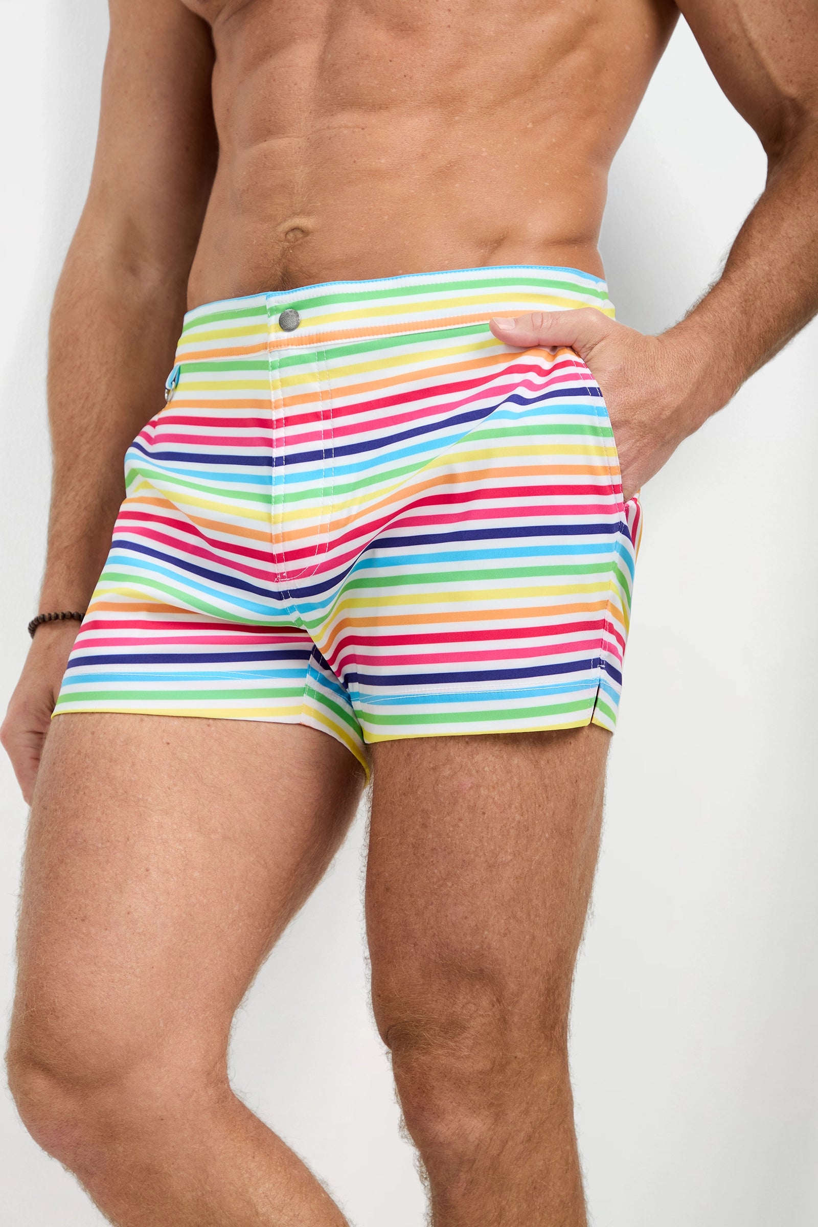 A muscular man models the Chico Swim Trunk - Pride, featuring bold horizontal stripes in red, blue, green, and yellow. He poses casually with one hand in his pocket against a plain background.