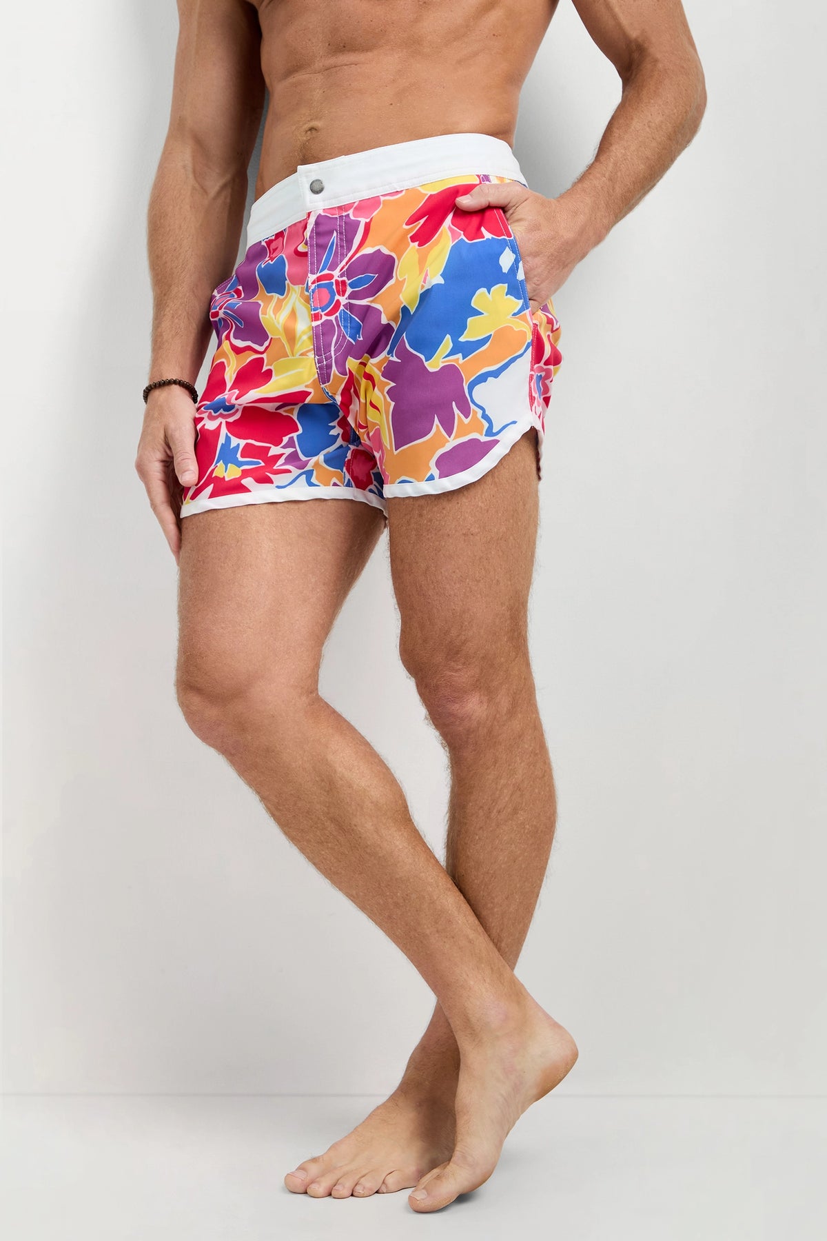 A person stands barefoot wearing the Surfside Board Short - Soie Paradis, featuring colorful floral patterns, a white waistband, and side split hems; their upper body is not fully visible.