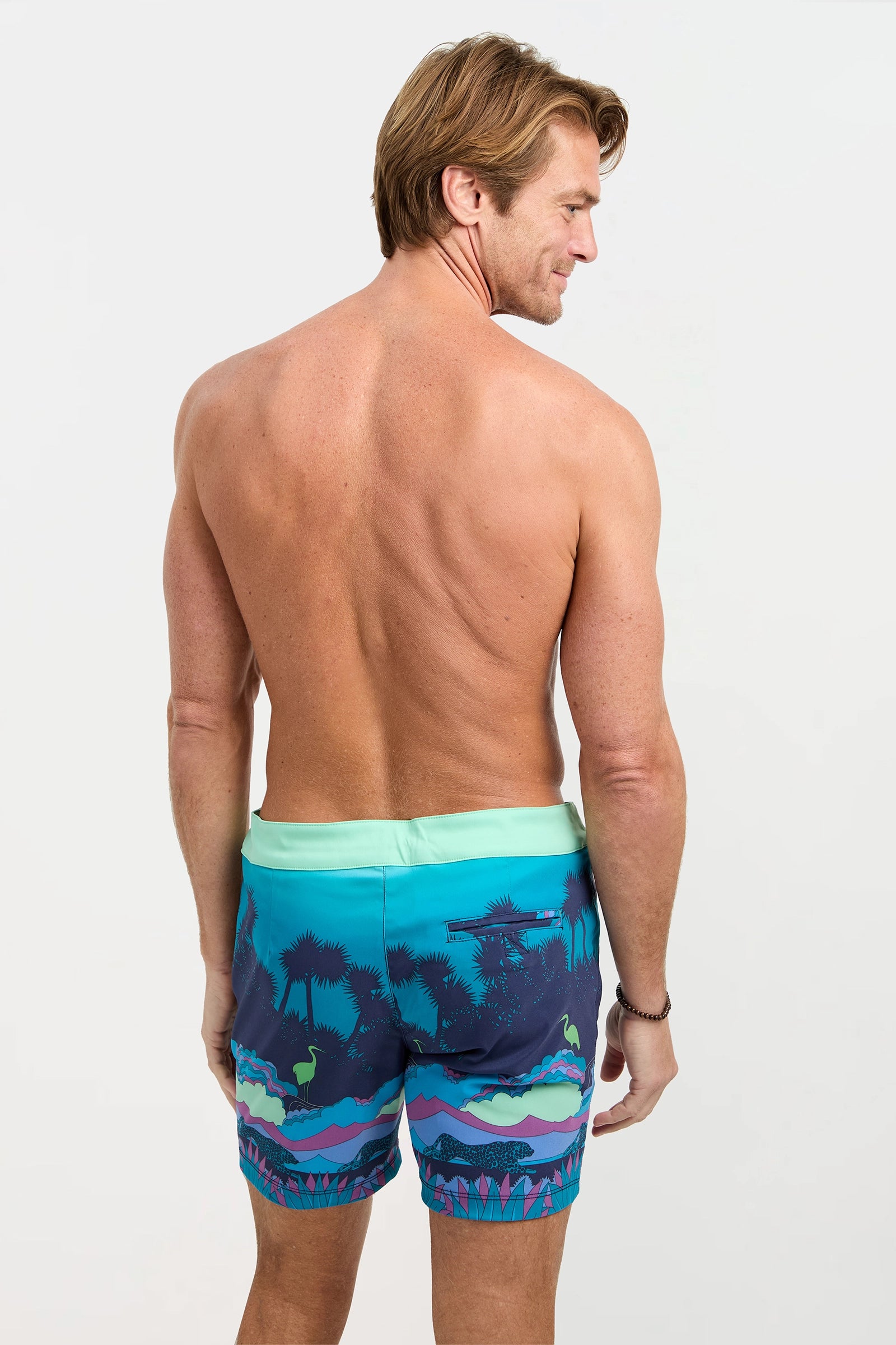 A man with light brown hair faces away, showing his bare back while wearing Honolulu Board Short - Jungle, featuring a colorful jungle swim print in blue, green, and purple tones.