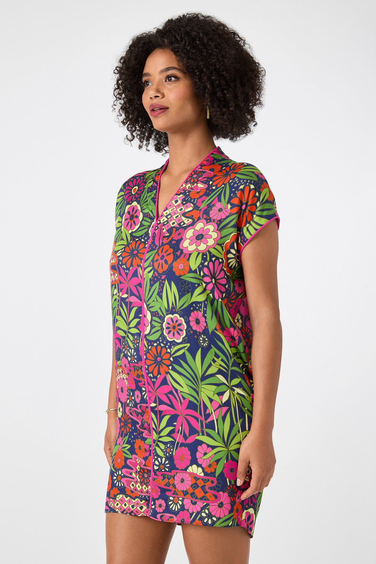 A woman with curly hair wears the Tarashi Short-Sleeve Mini Dress - Fumi Floral, showcasing vibrant pink, green, orange, and purple flowers and leaves, standing against a plain white background.