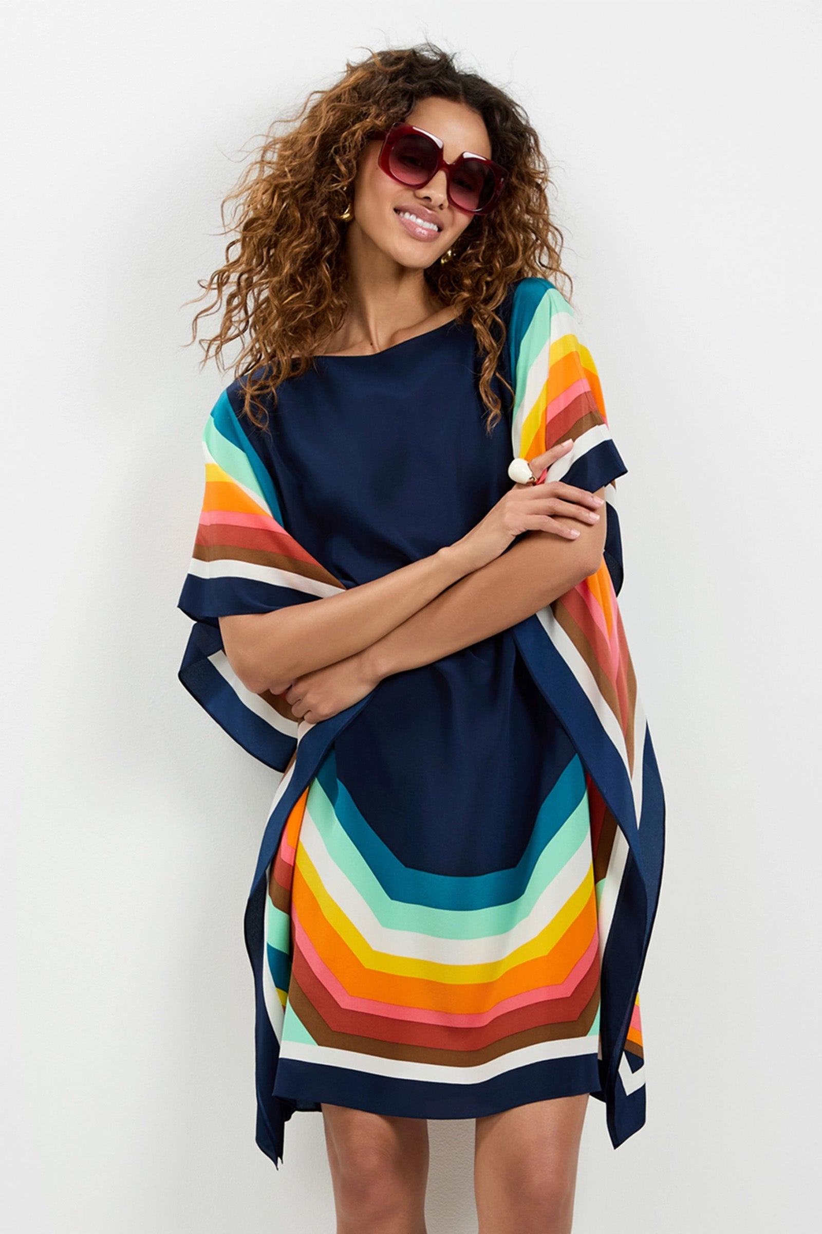 A woman with curly hair and sunglasses smiles with her arms crossed, wearing the Theodora Caftan Dress - Siena, which features colorful horizontal stripes and a navy blue base, against a plain white background.
