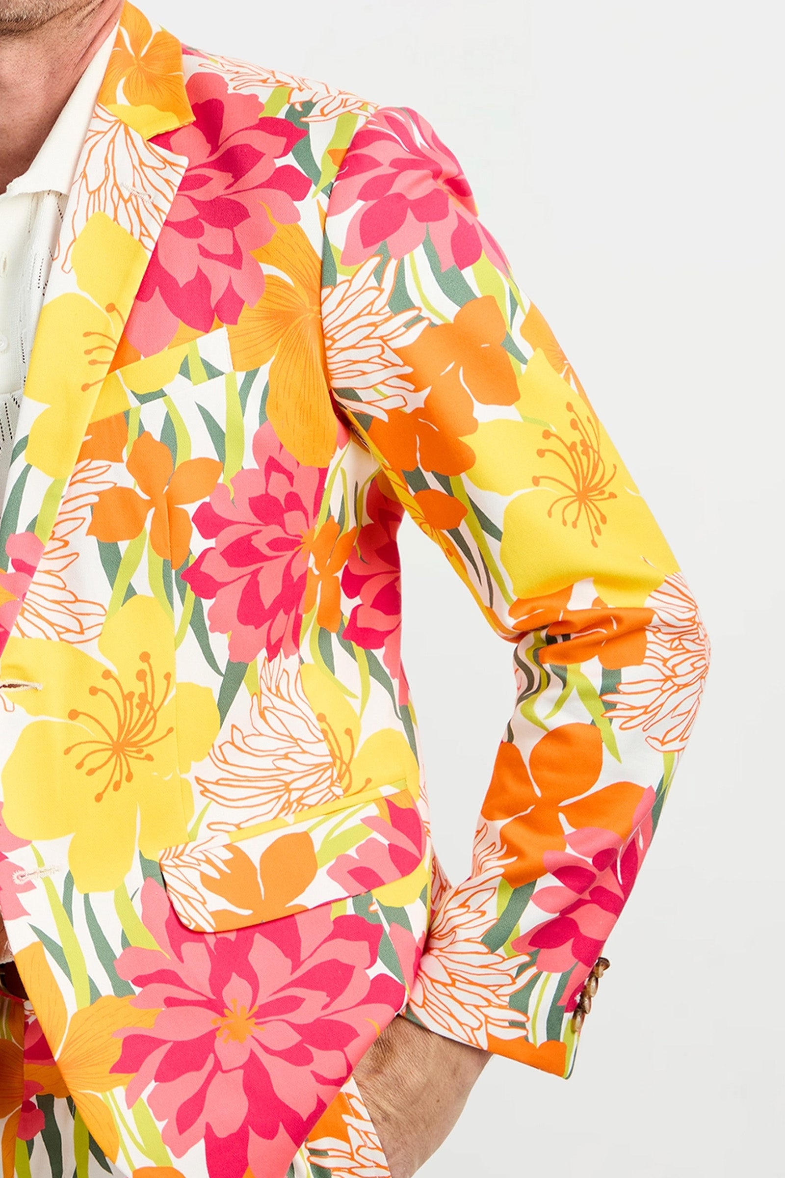 A person wearing the Thurston Blazer - Como Herringbone, featuring a bold floral pattern in yellow, orange, pink, and white, stands against a plain light background. Only the left half of their upper body is visible.