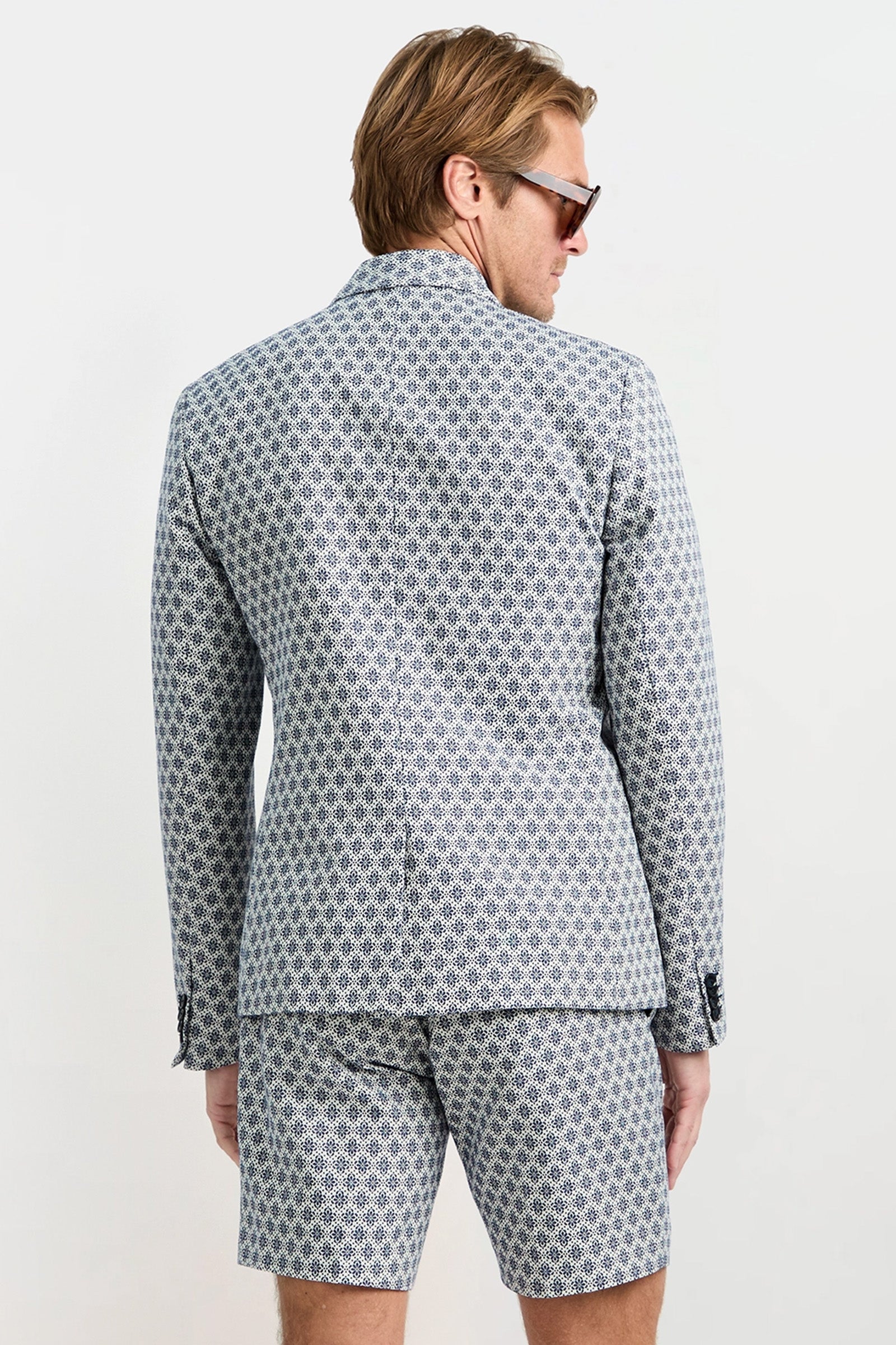 A man with light brown hair and sunglasses stands with his back to the camera against a plain white background, wearing the Thurston Blazer - Daisy Jacquard and matching shorts.