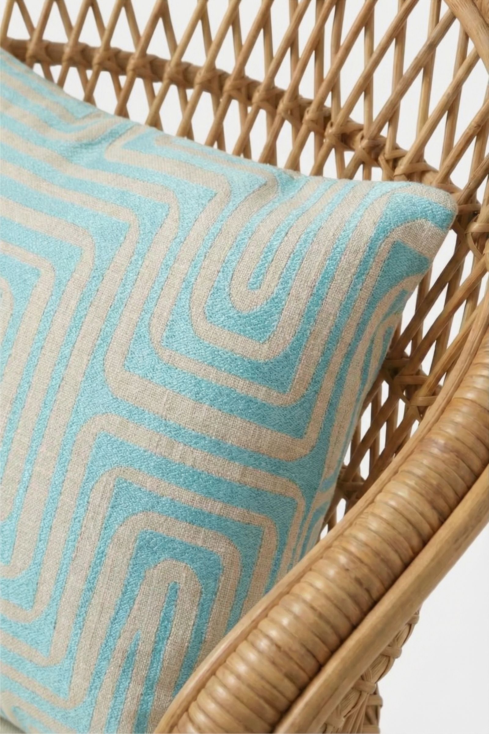 A close-up of the Amazing Maze Embroidered Pillow, featuring a light blue and beige geometric pattern, placed on a woven rattan chair against a white background.