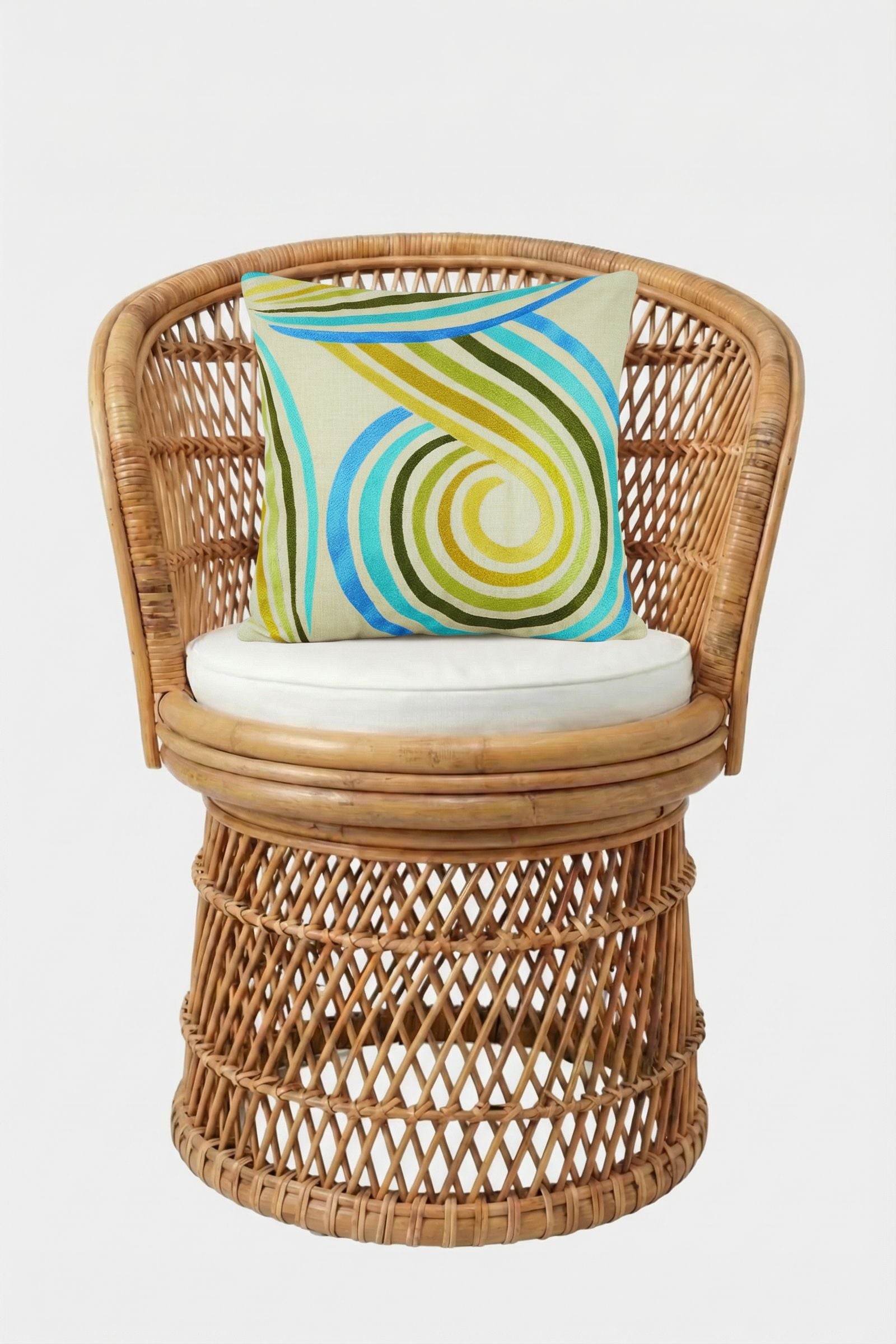 A round wicker chair with a white cushion holds the CALEXICO EMB PILLOW 20X20", which features a colorful blue, green, and yellow spiral pattern and plush down feather fill, all set against a plain white background.