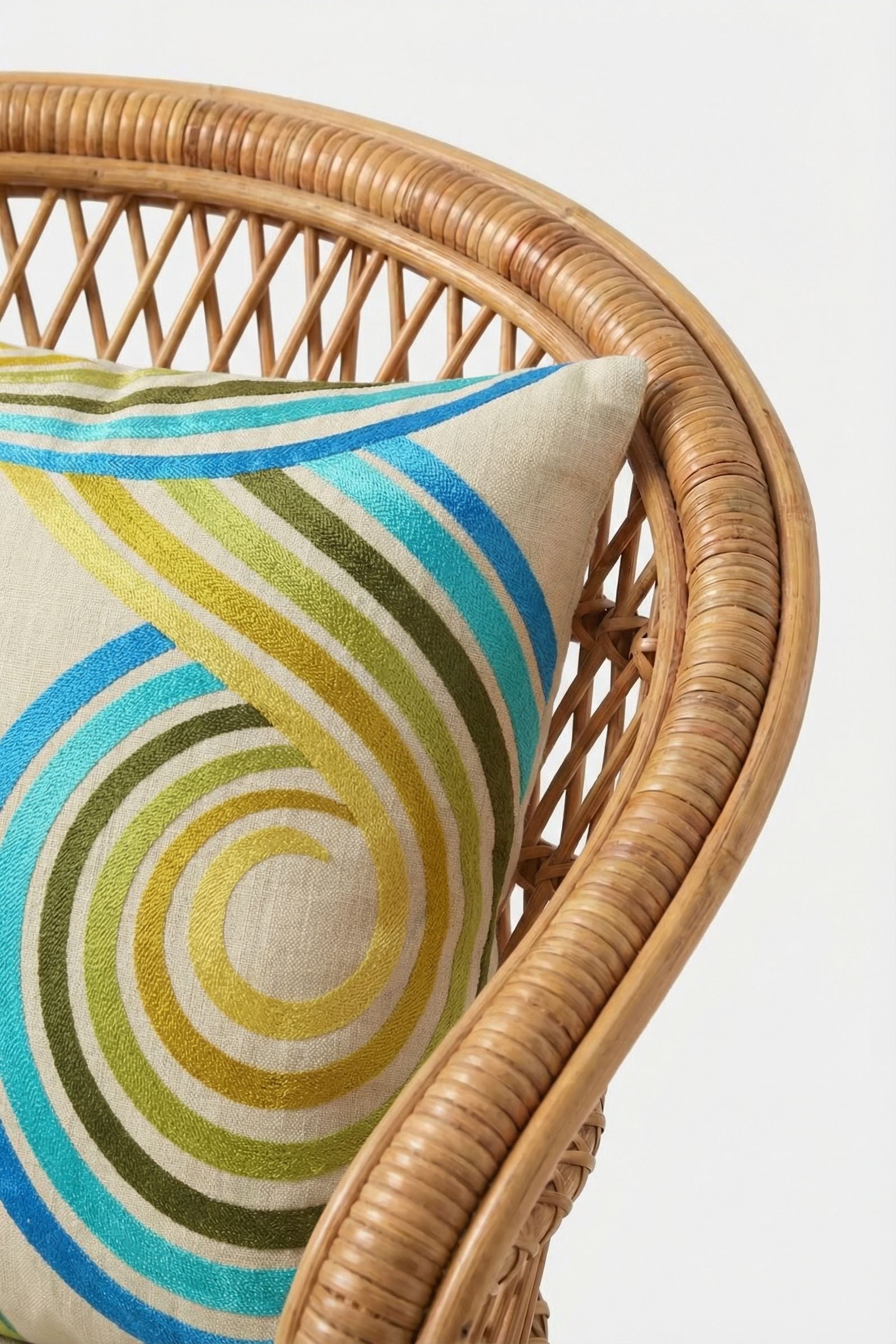 A close-up of a rattan chair with the CALEXICO EMB PILLOW 20X20", featuring blue, green, and yellow swirling lines on a light background. The image showcases the curved arm and the vibrant down-filled cushion.